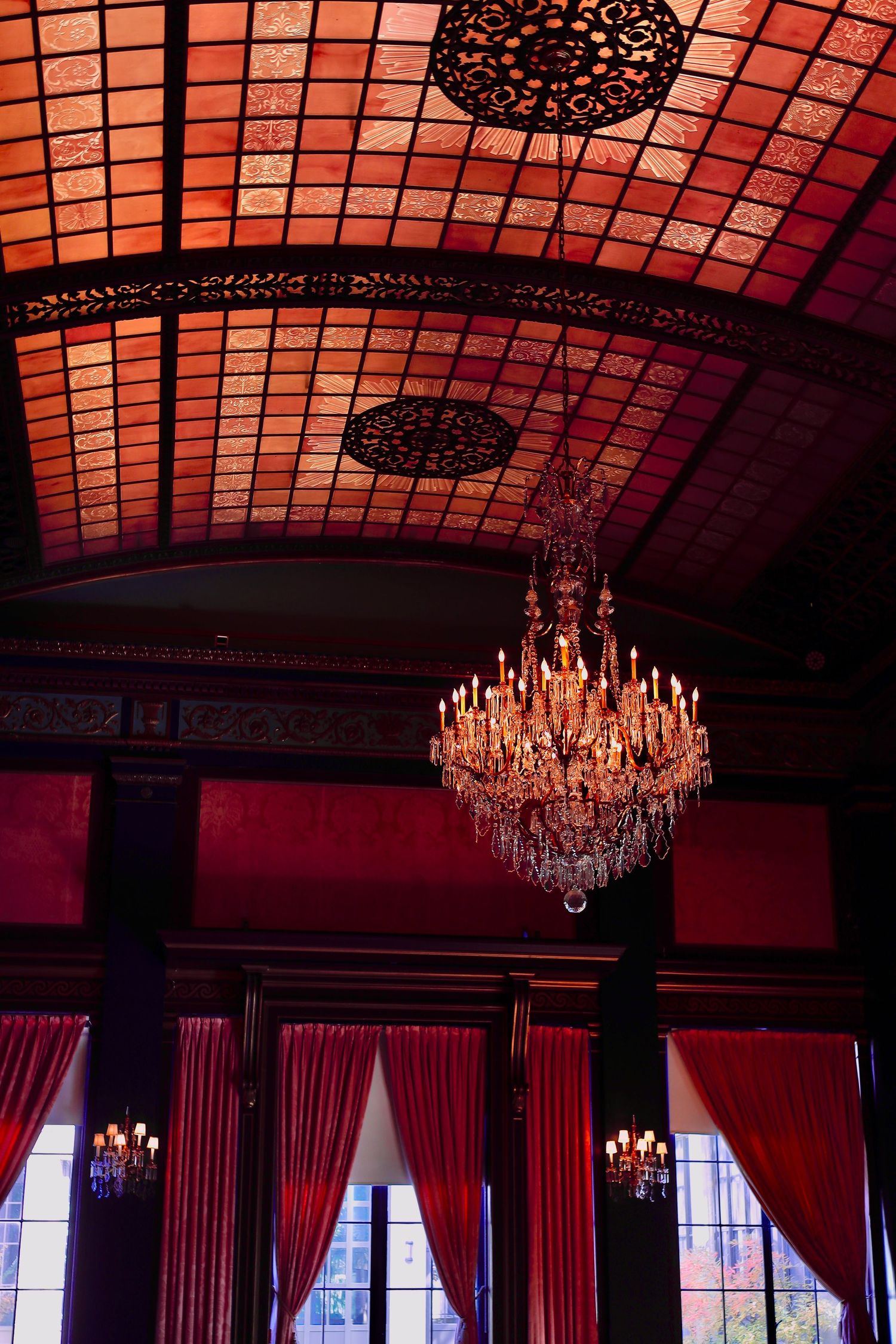 Ornate crystal chandelier hangs in grand ballroom with red walls, dramatic curtains and tiled glass ceiling.