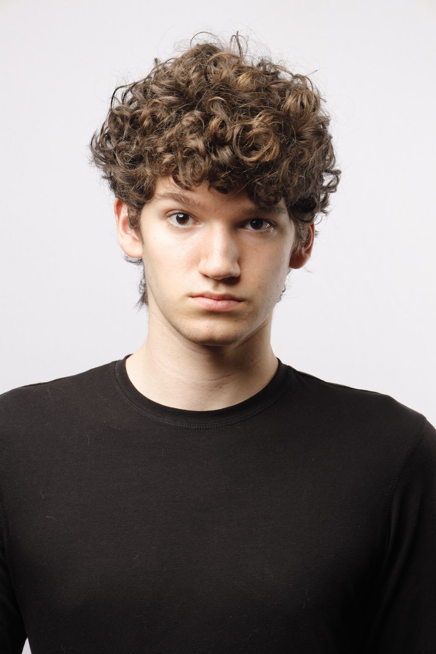A person with curly brown hair wearing a black shirt poses against a white backdrop for a composition photo.