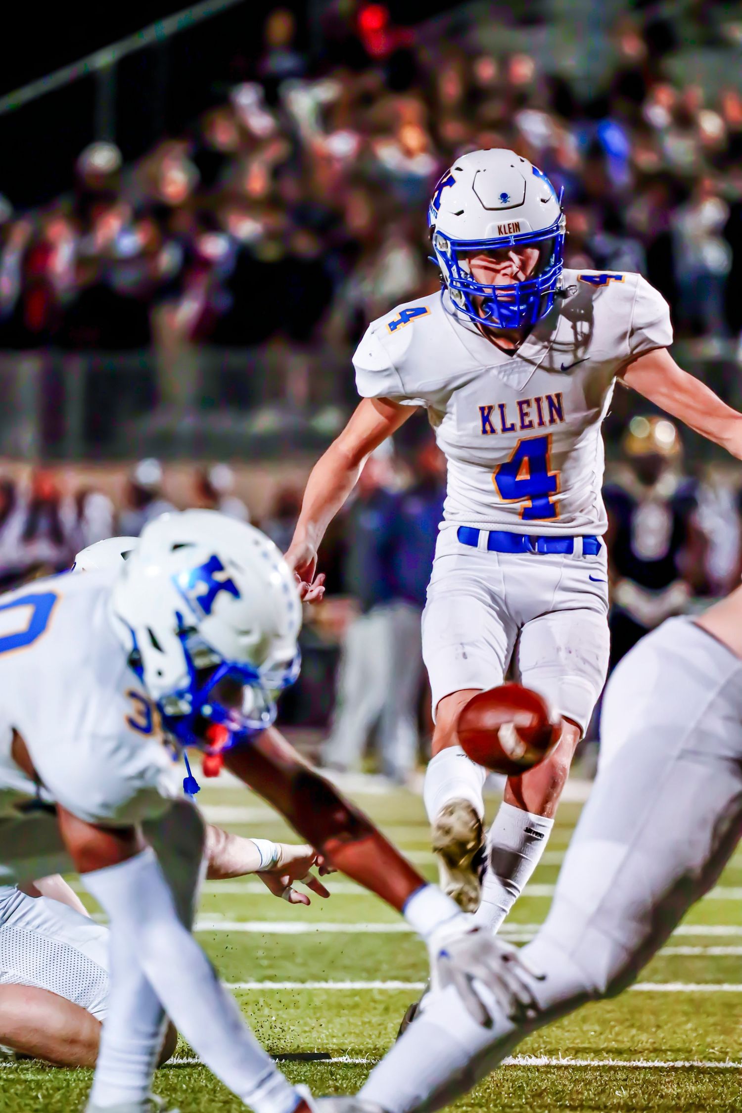 High school football player in white Klein uniform kicks field goal during night game while teammate holds ball.