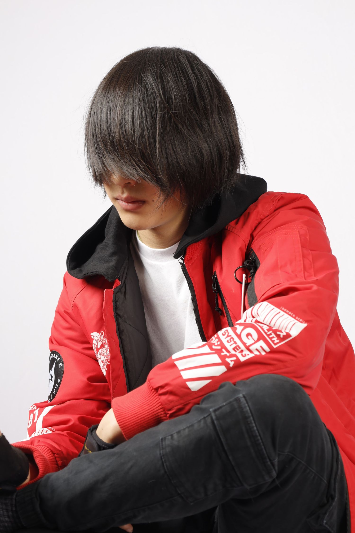 Person wearing red North Face jacket with dark hair and white t-shirt sits in casual pose against white background.