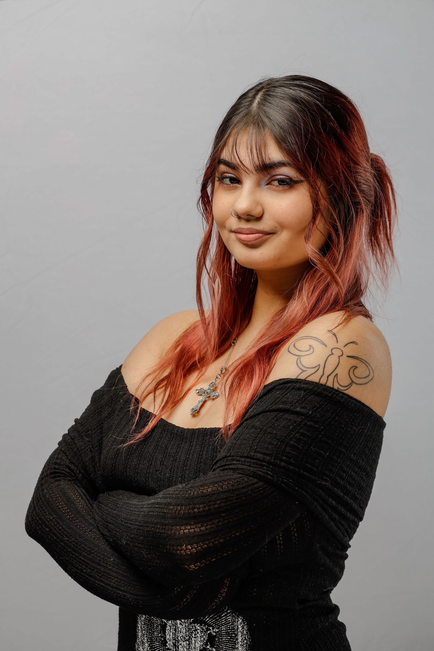 Portrait of someone with pink ombre hair wearing a black off-shoulder sweater posing against a gray background.