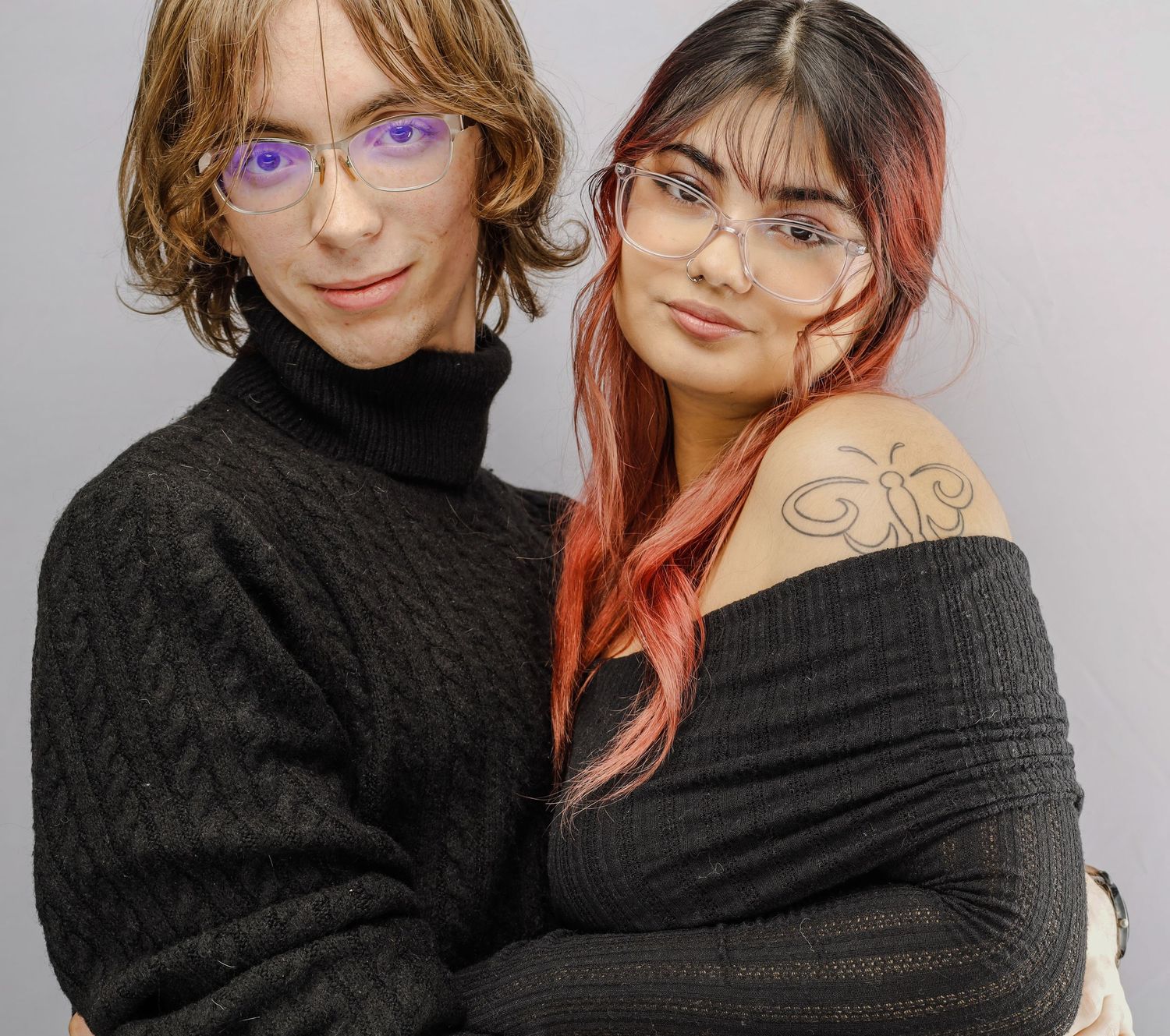 Two people wearing black sweaters pose together, one with purple glasses and another with dark rimmed glasses and pink hair.