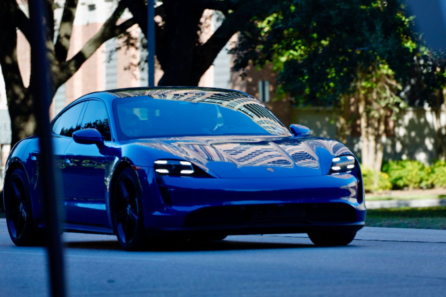 Electric blue Porsche Taycan sports car parked under tree shadows.