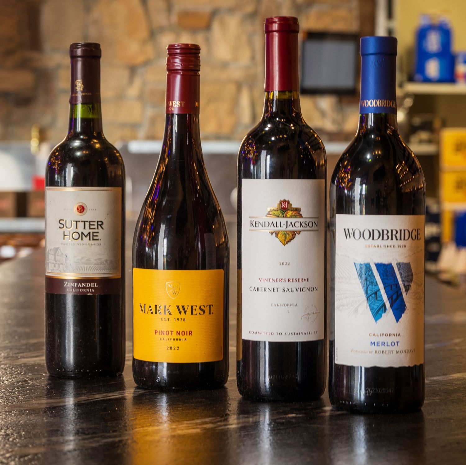 Four bottles of red wine including Sutter Home, Nine West, Kendall Jackson, and Woodbridge displayed on a wooden surface.