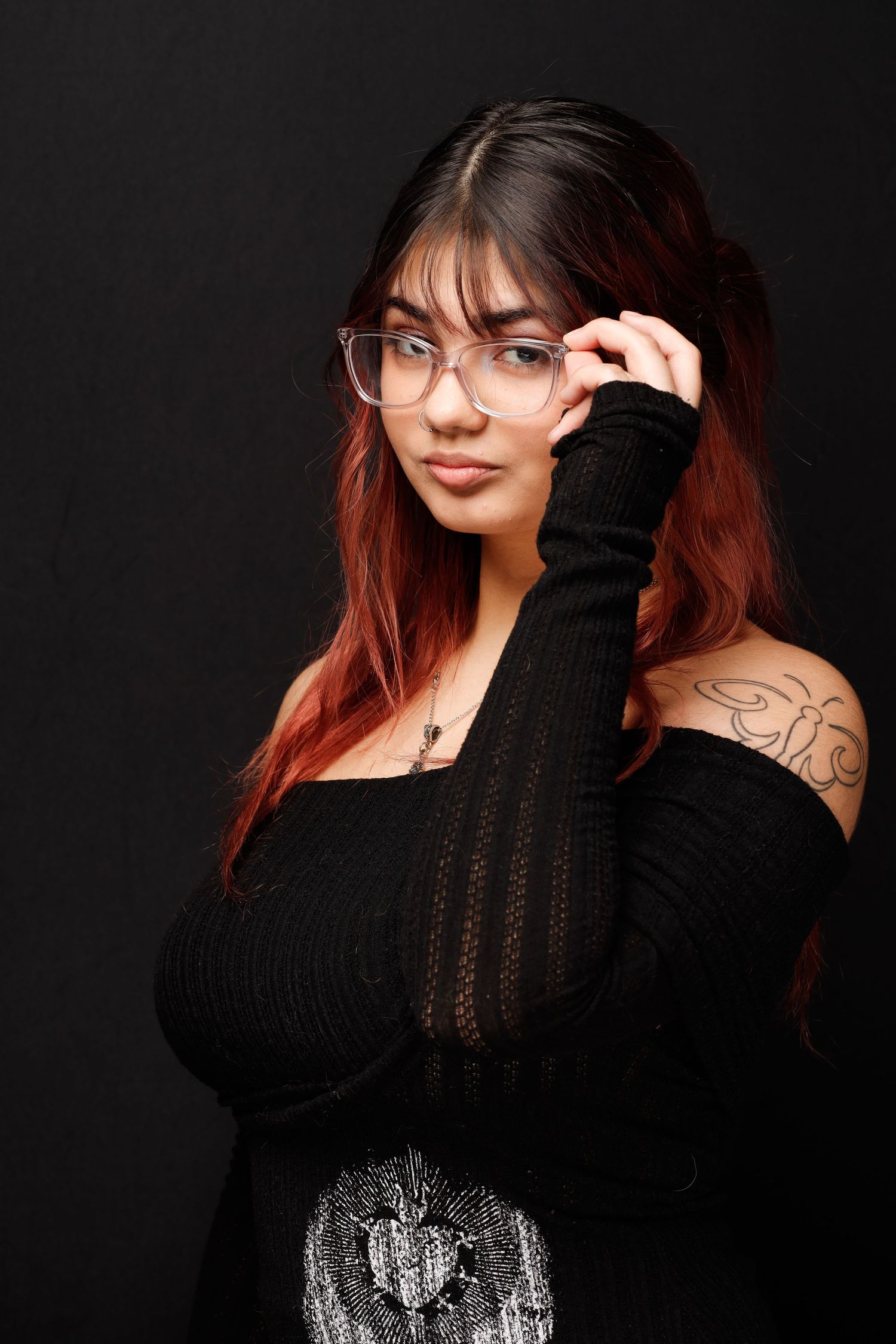 Portrait of someone with long red-black ombre hair wearing glasses and a black sweater against dark background.