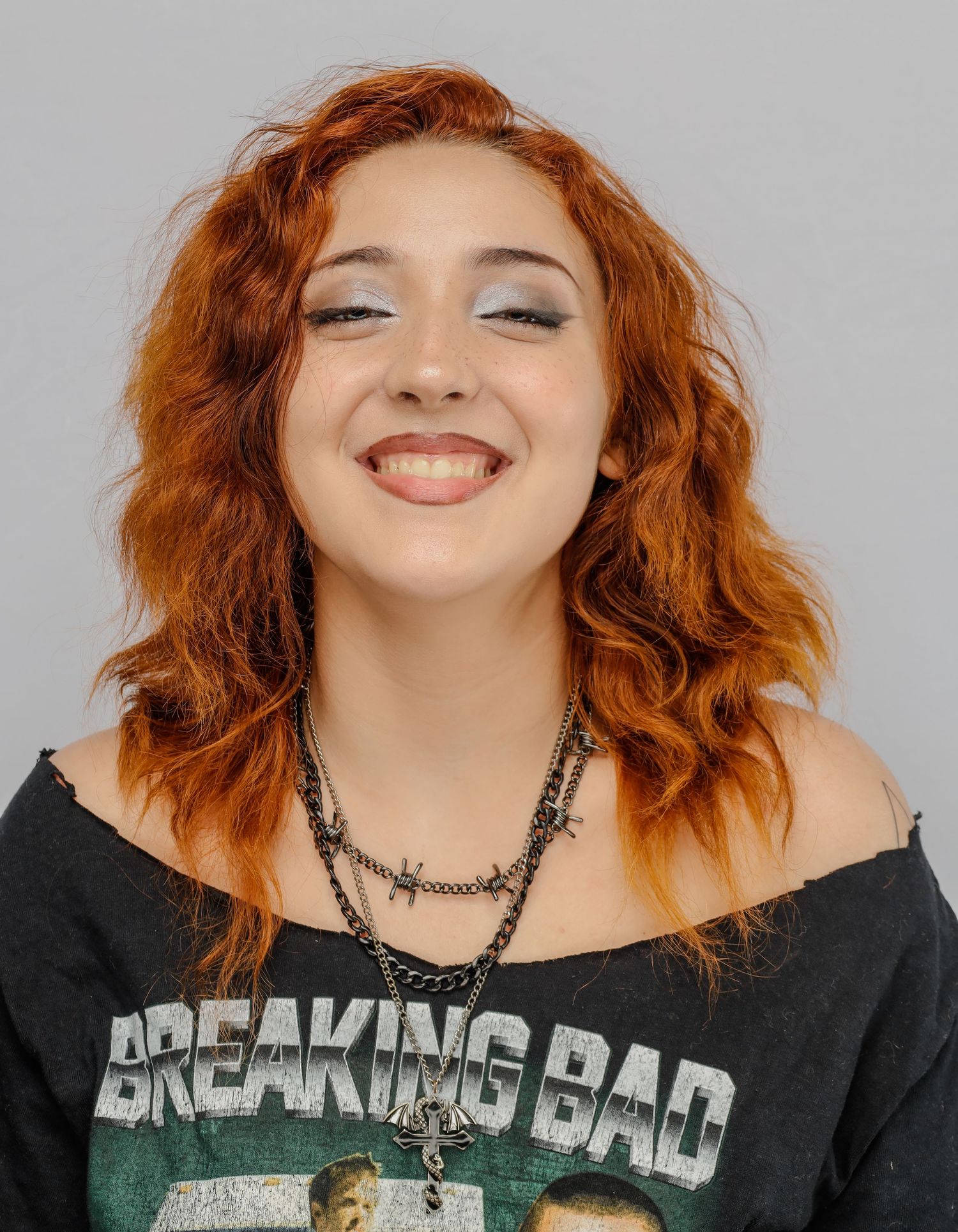 A smiling person with curly red hair wearing an off-shoulder Breaking Bad t-shirt and dark necklace.