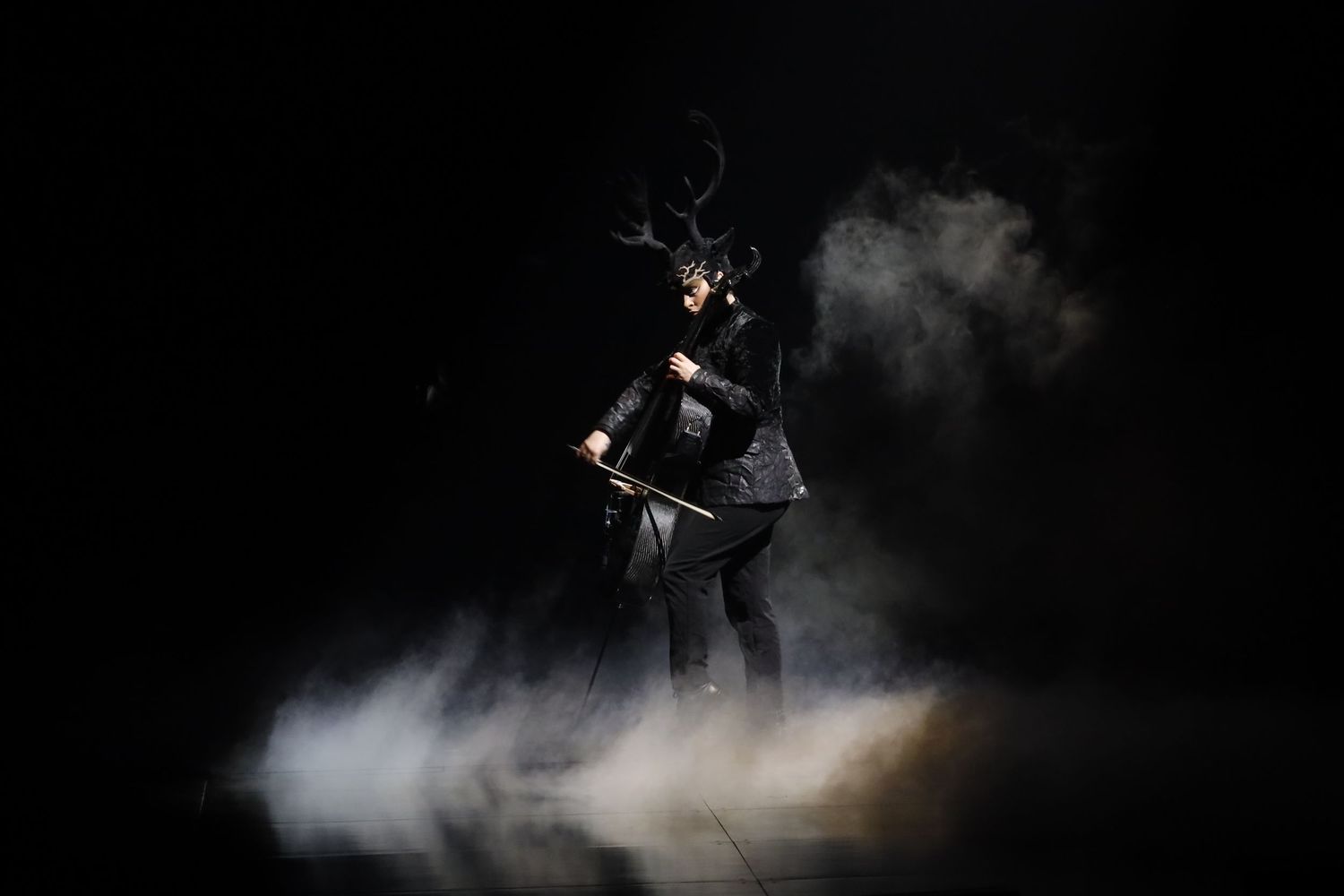 Mysterious figure in antlers and dark clothing emerges from smoke against black background.
