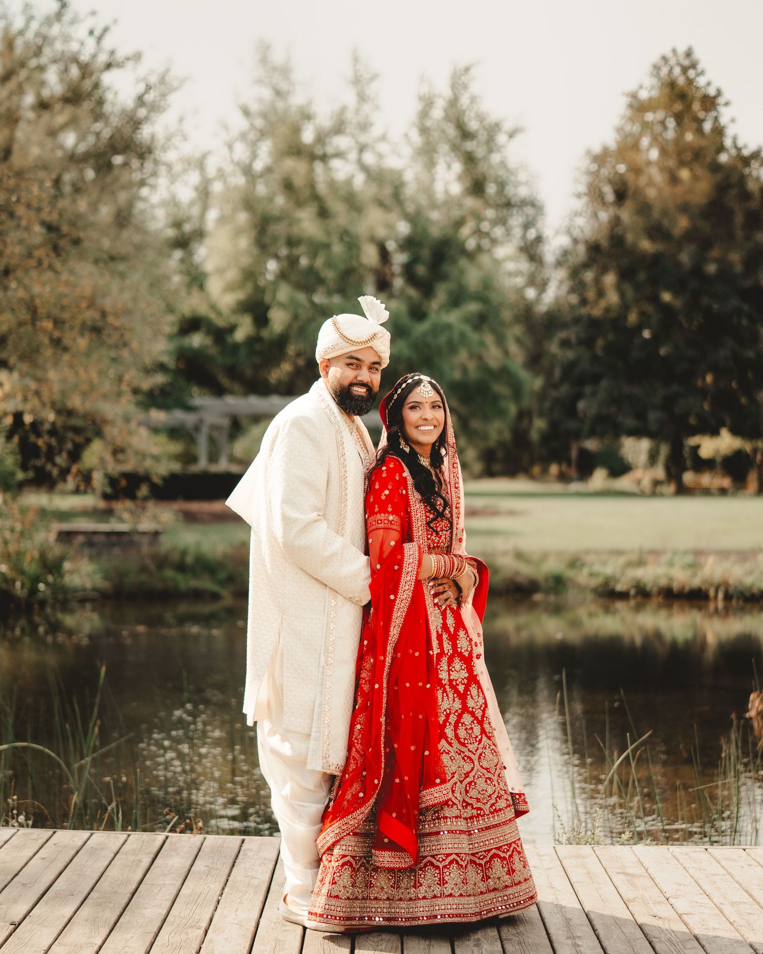 A couple in traditional Indian wedding attire stands on a wooden deck near a lake in autumn.