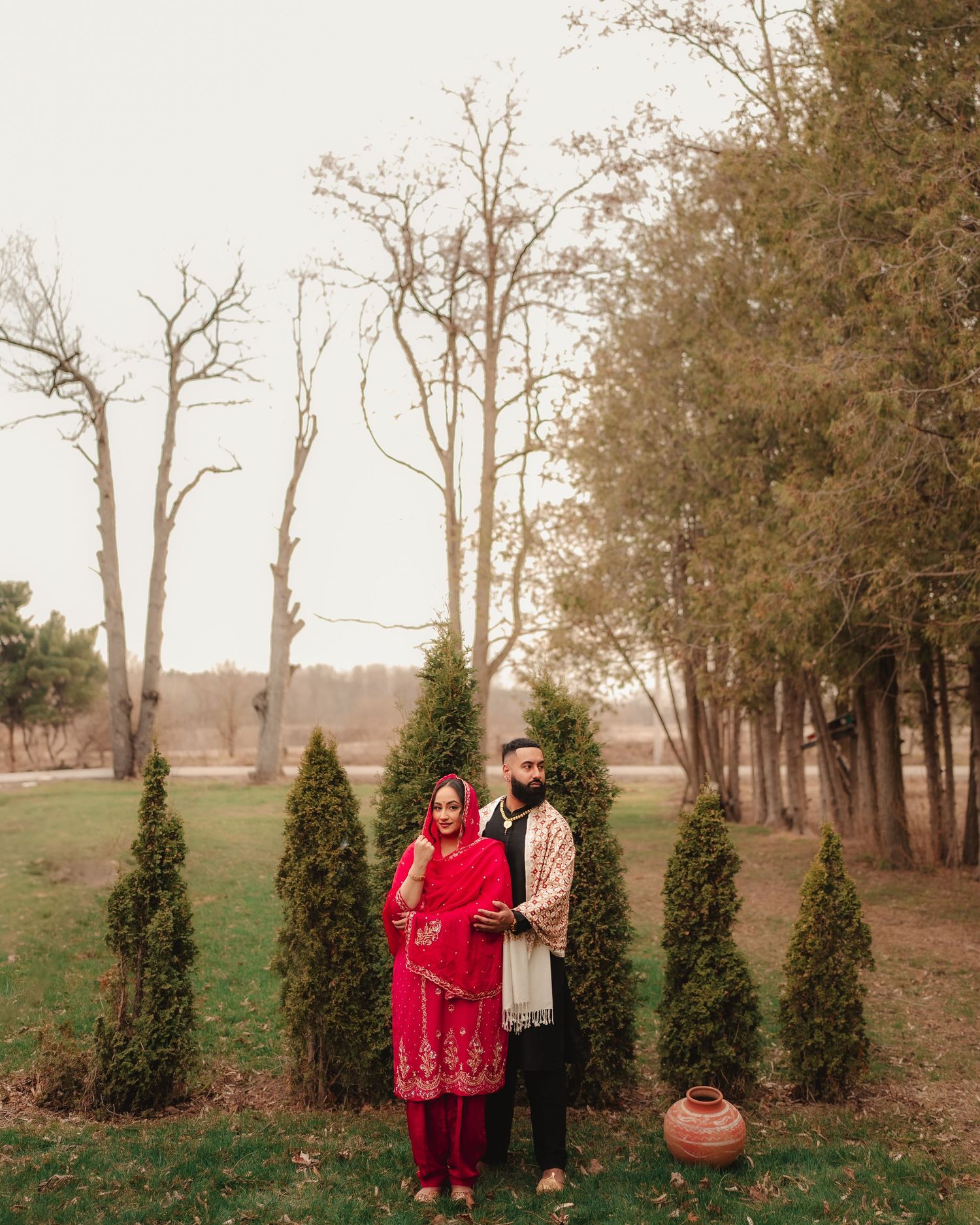 A couple stands among evergreen trees in a rustic outdoor setting during golden hour.