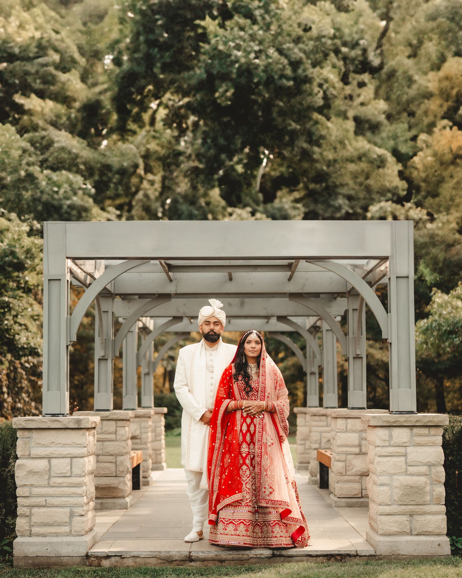 A couple in traditional Indian wedding dress poses under an elegant stone archway.