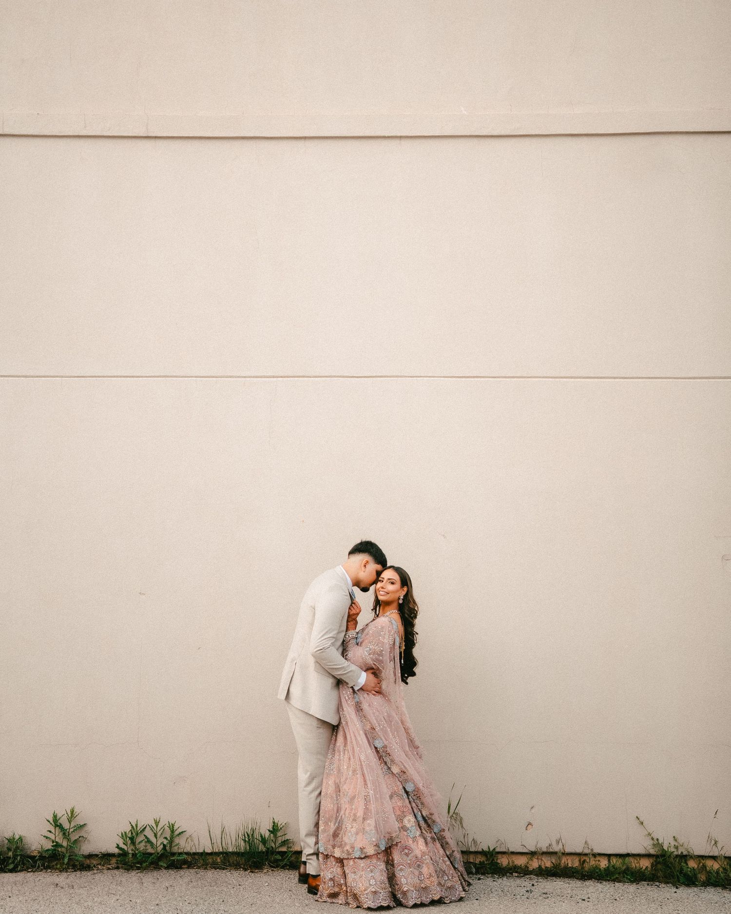A couple in formal attire embrace against a beige wall, with the woman wearing an elegant pink lehenga.