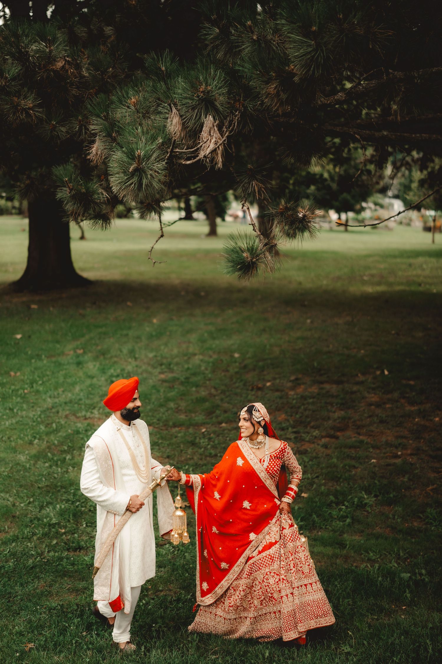 Couple in Sikh wedding attire walking together on green grass under pine trees.