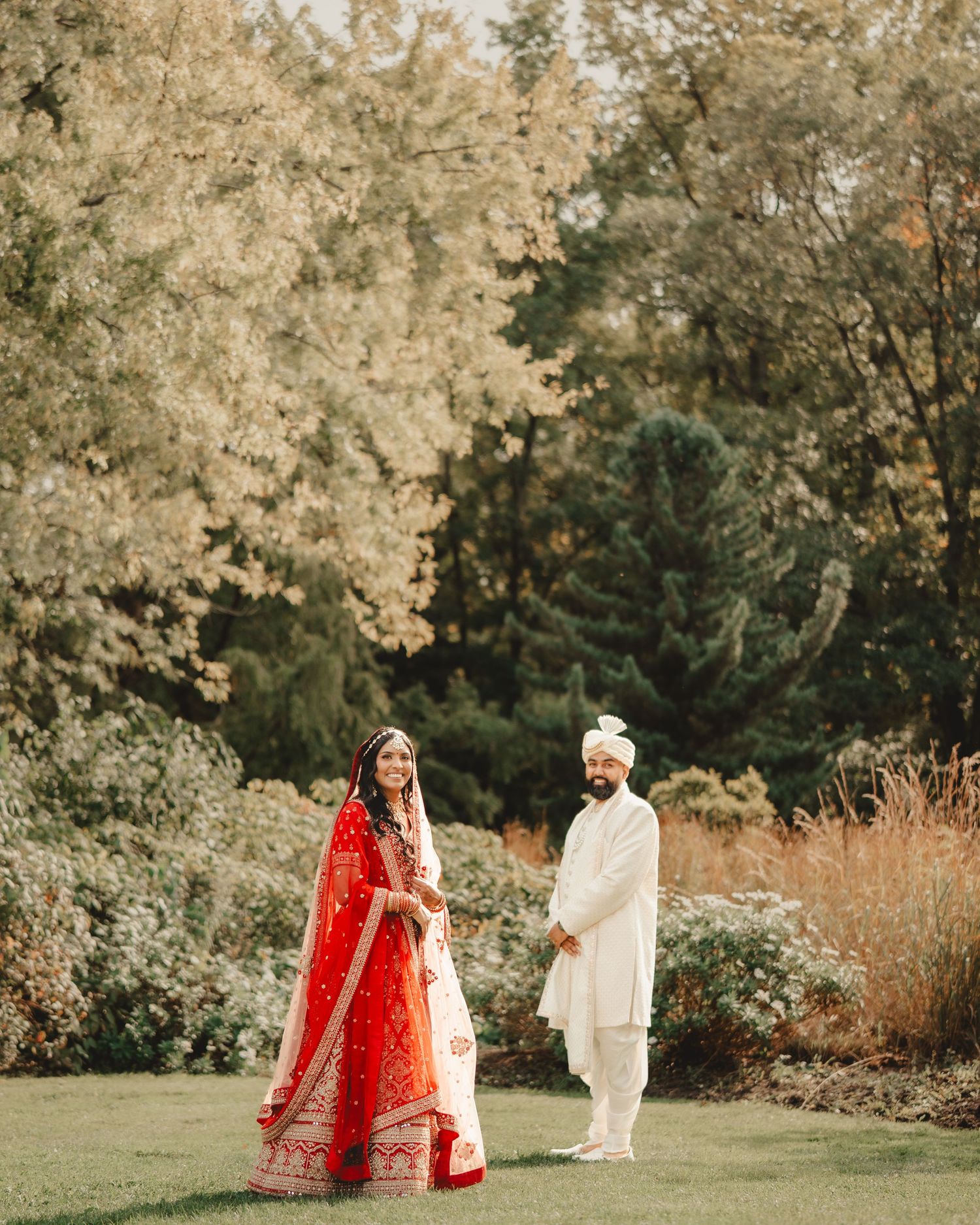 A couple in traditional Indian wedding attire poses in a garden setting with autumn trees.