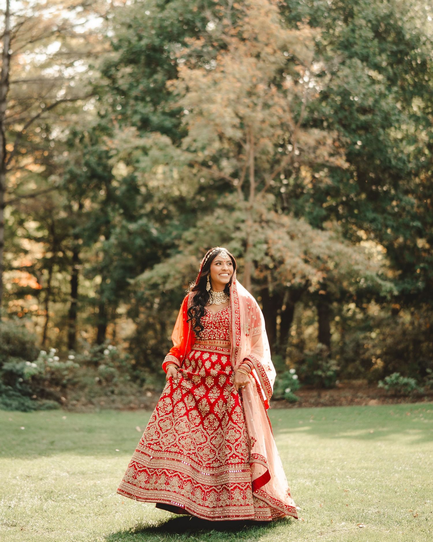 A bride in a red lehenga twirls in a garden setting surrounded by autumn trees.