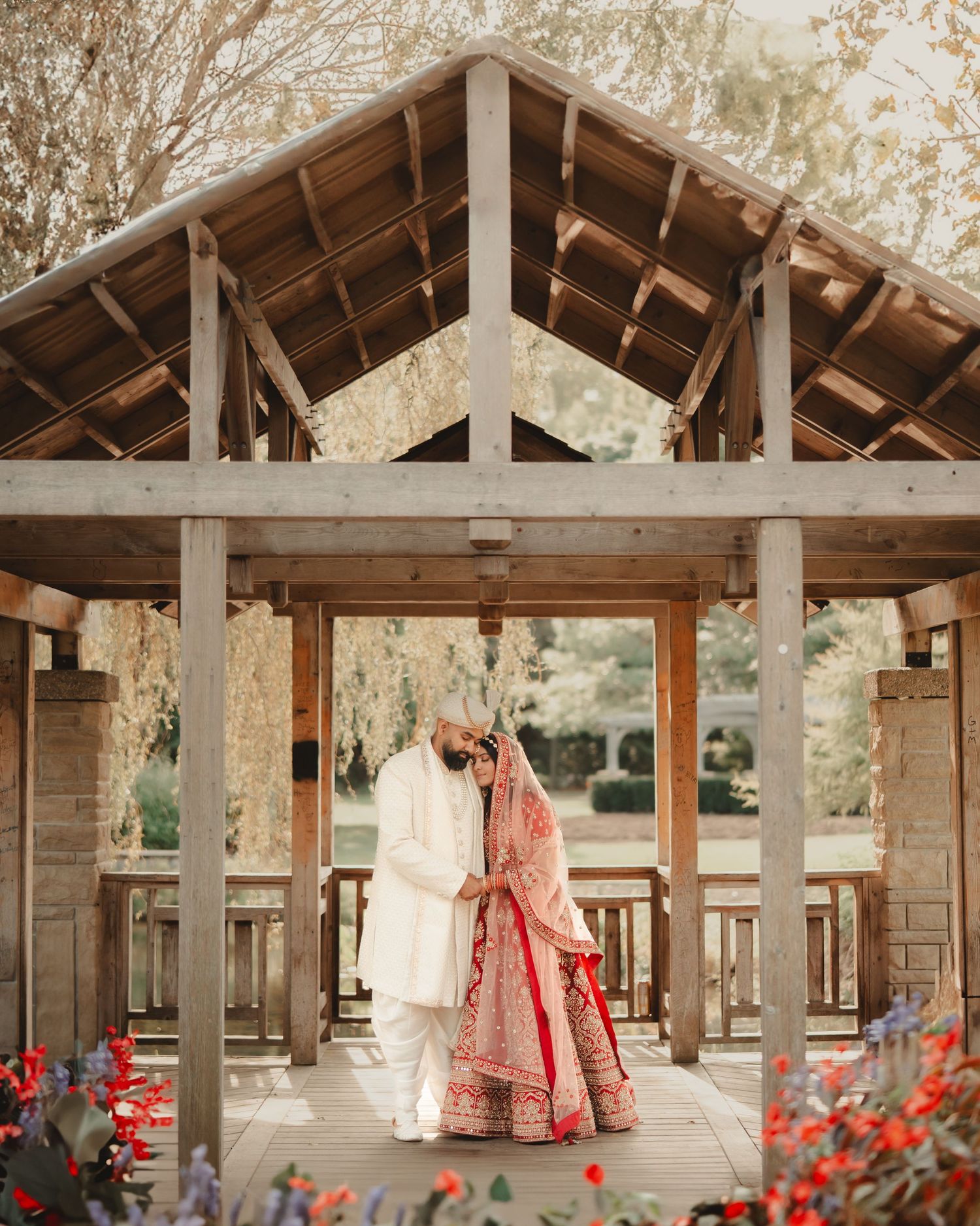 A couple in traditional Indian wedding dress stands beneath a wooden pavilion with flowers.