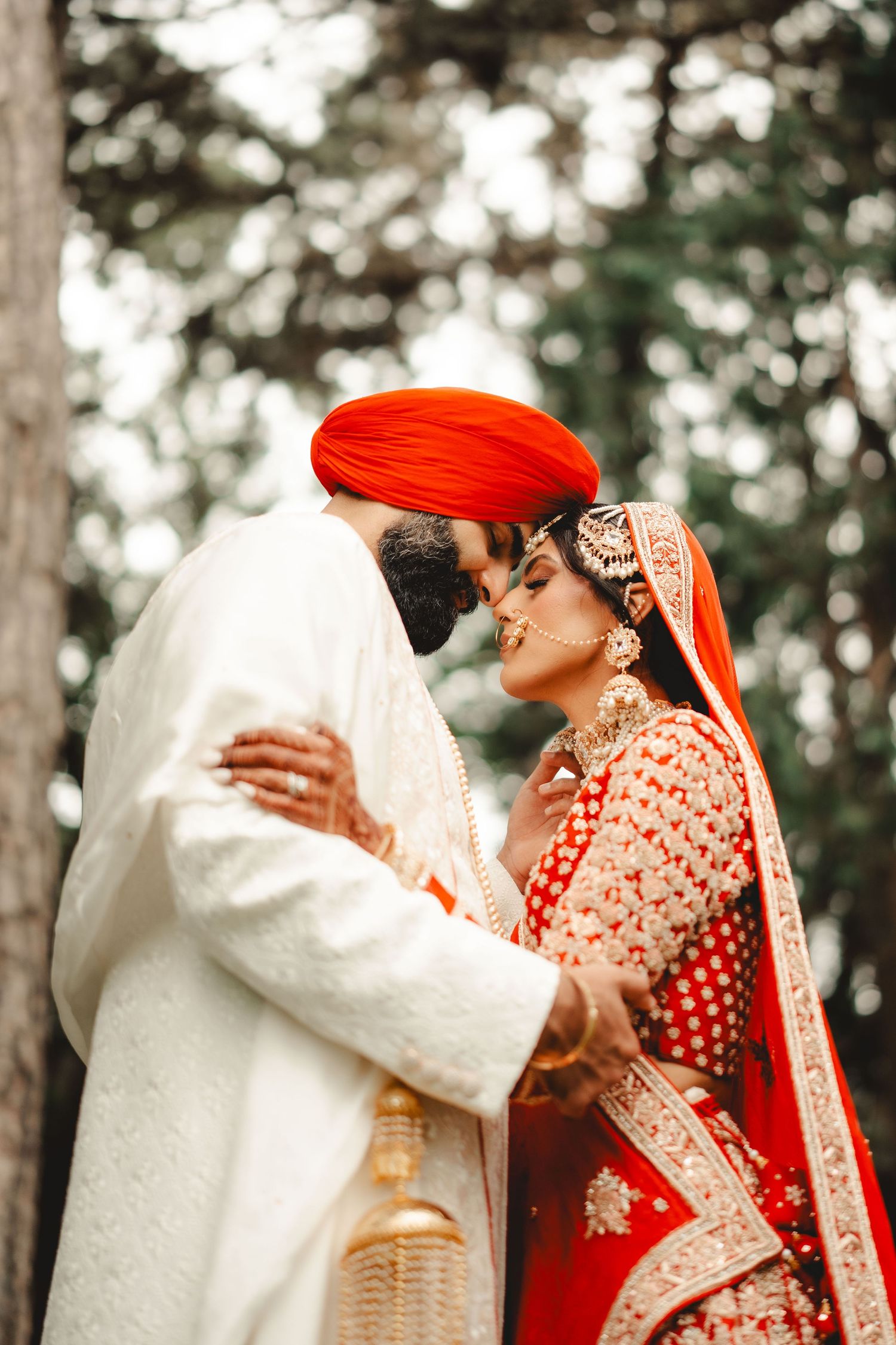 Intimate moment between newlyweds in traditional Sikh wedding attire amongst pine trees.