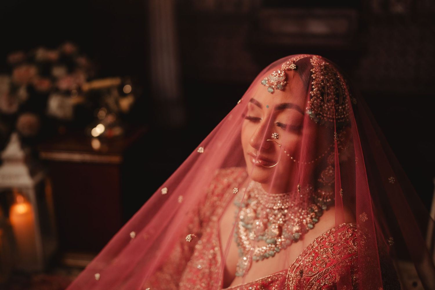 A bride in traditional South Asian wedding attire wears an ornate red veil and glittering jewelry in soft candlelight.
