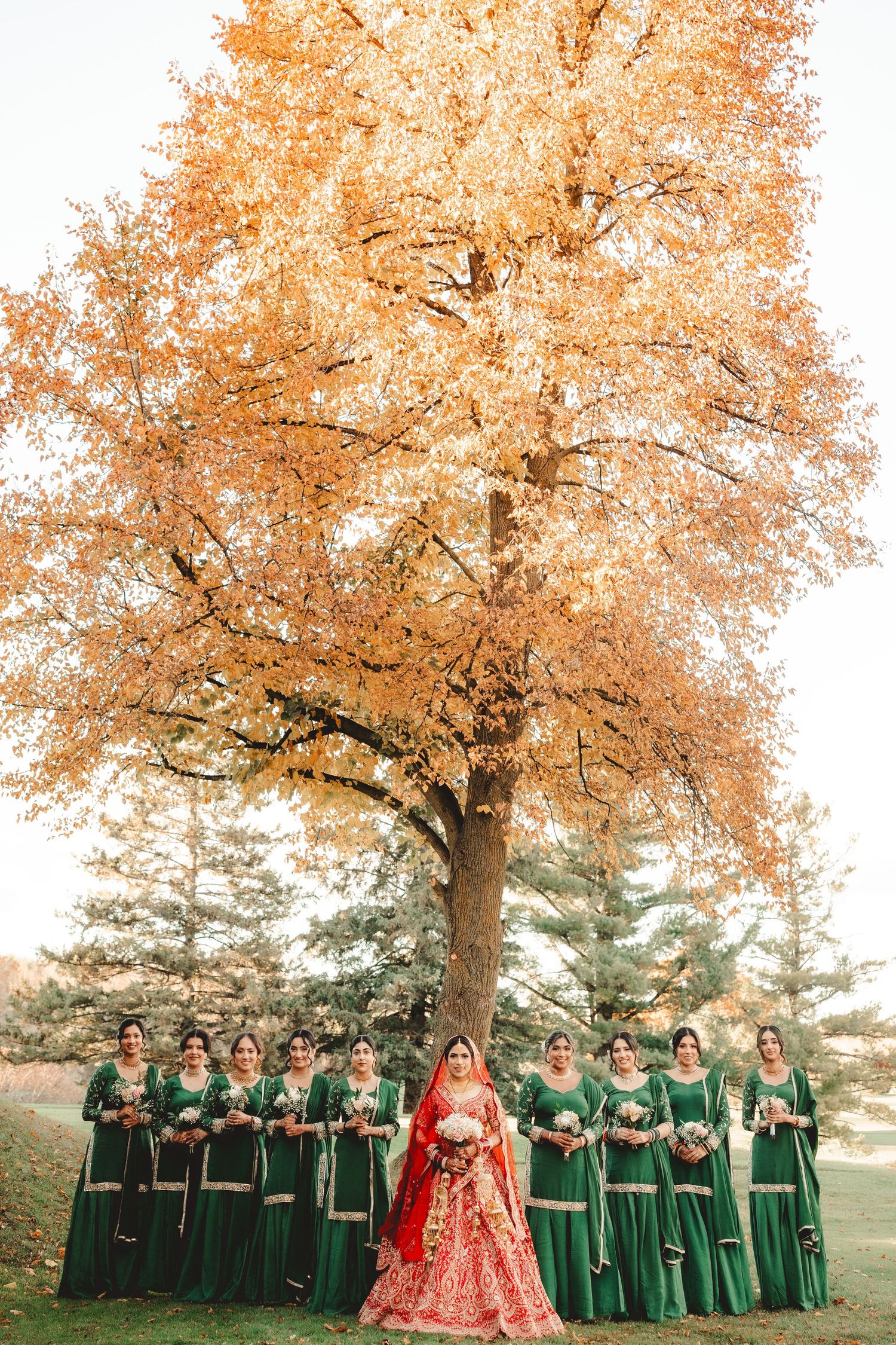 Wedding party in emerald green dresses poses with bride in red lehenga under autumn trees with orange foliage.