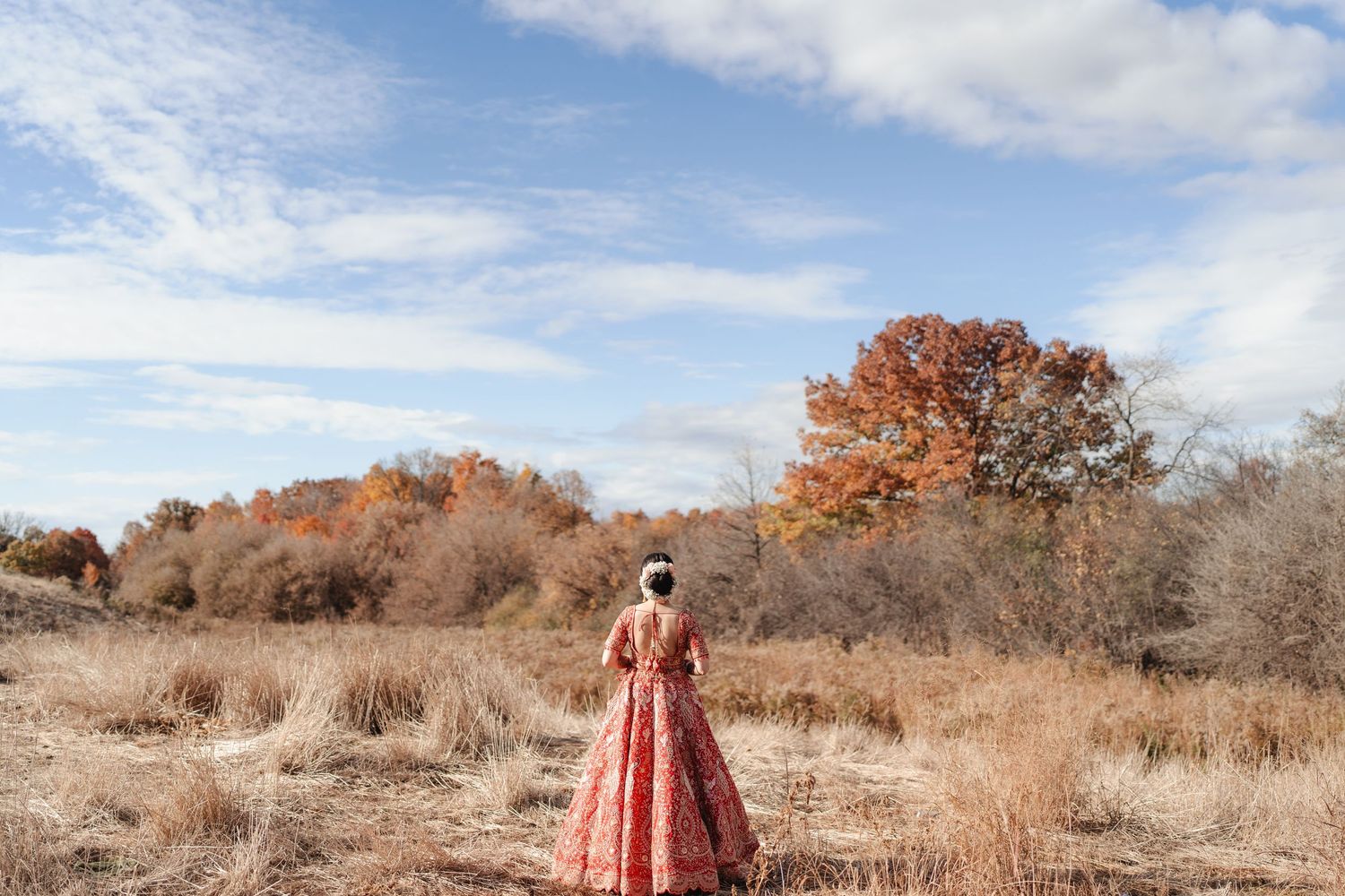 Person in red wedding attire standing in an autumn field with trees in the background.