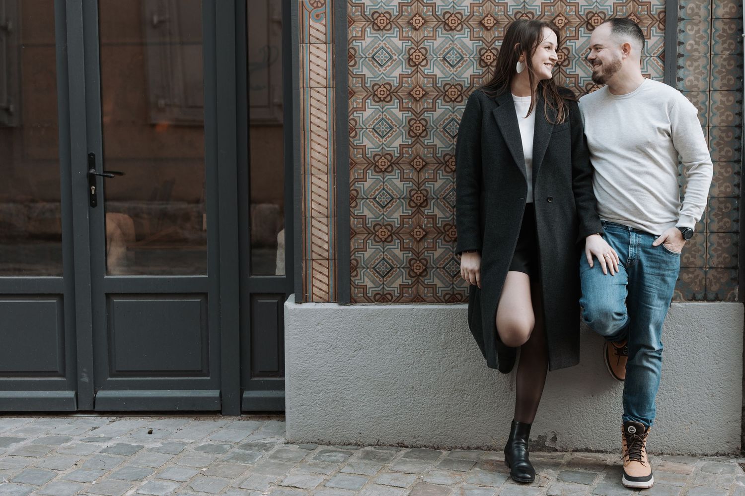 Stylish couple posing against vintage tiled wall with dark doorways.