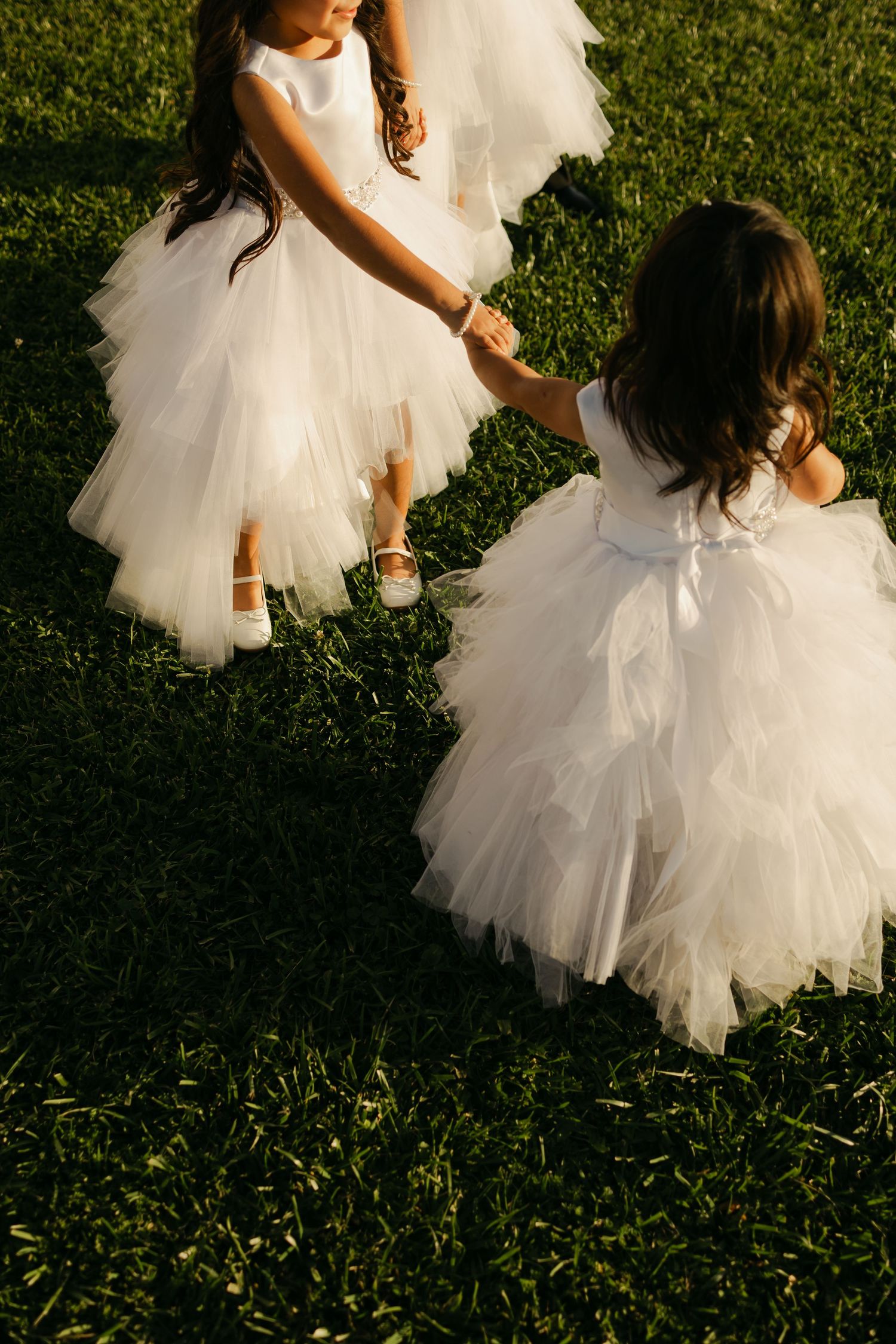Two flower girls in white tulle dresses playing on grass in golden sunlight.