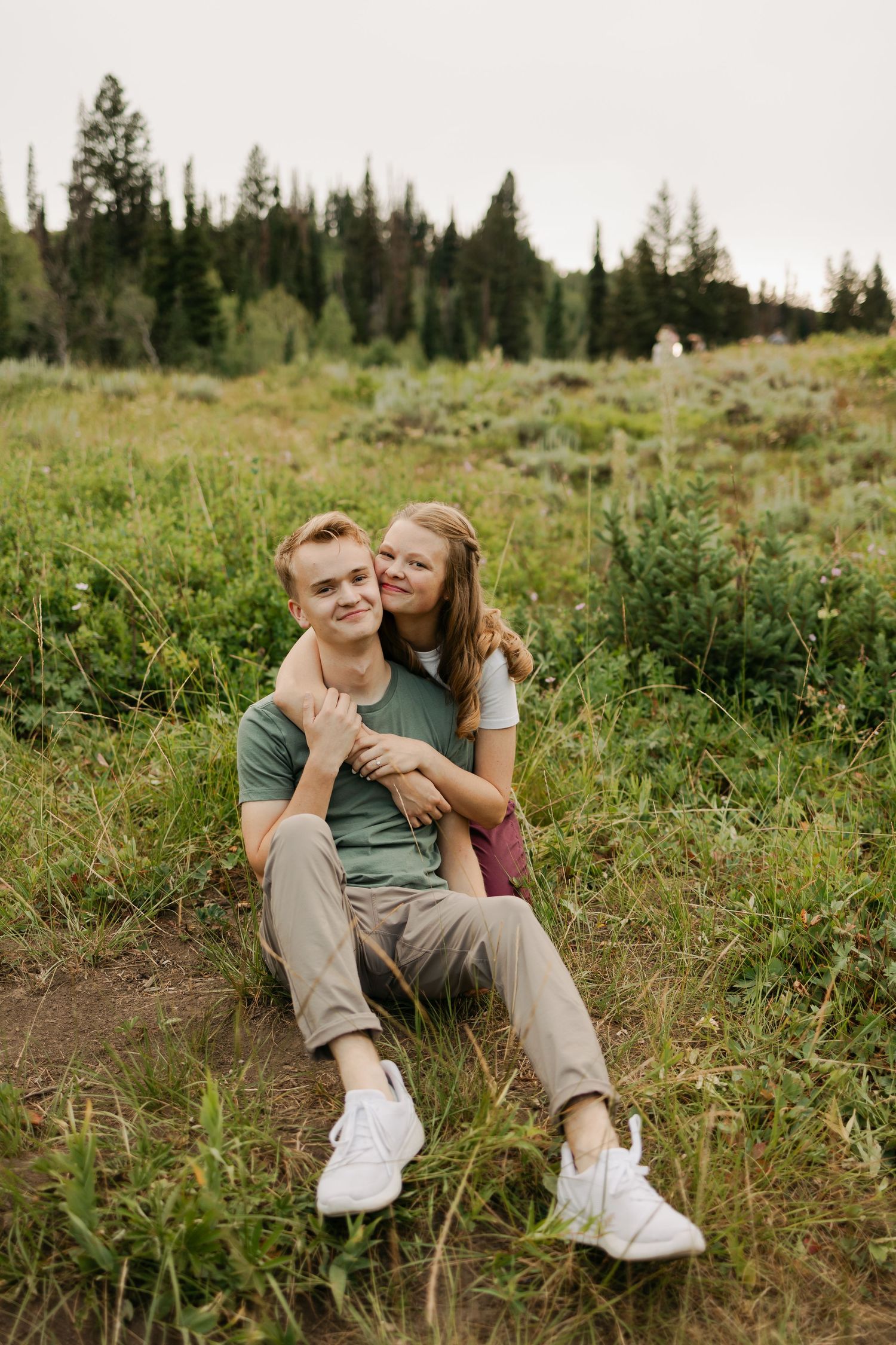A couple relaxes together on a grassy hillside surrounded by evergreen trees.