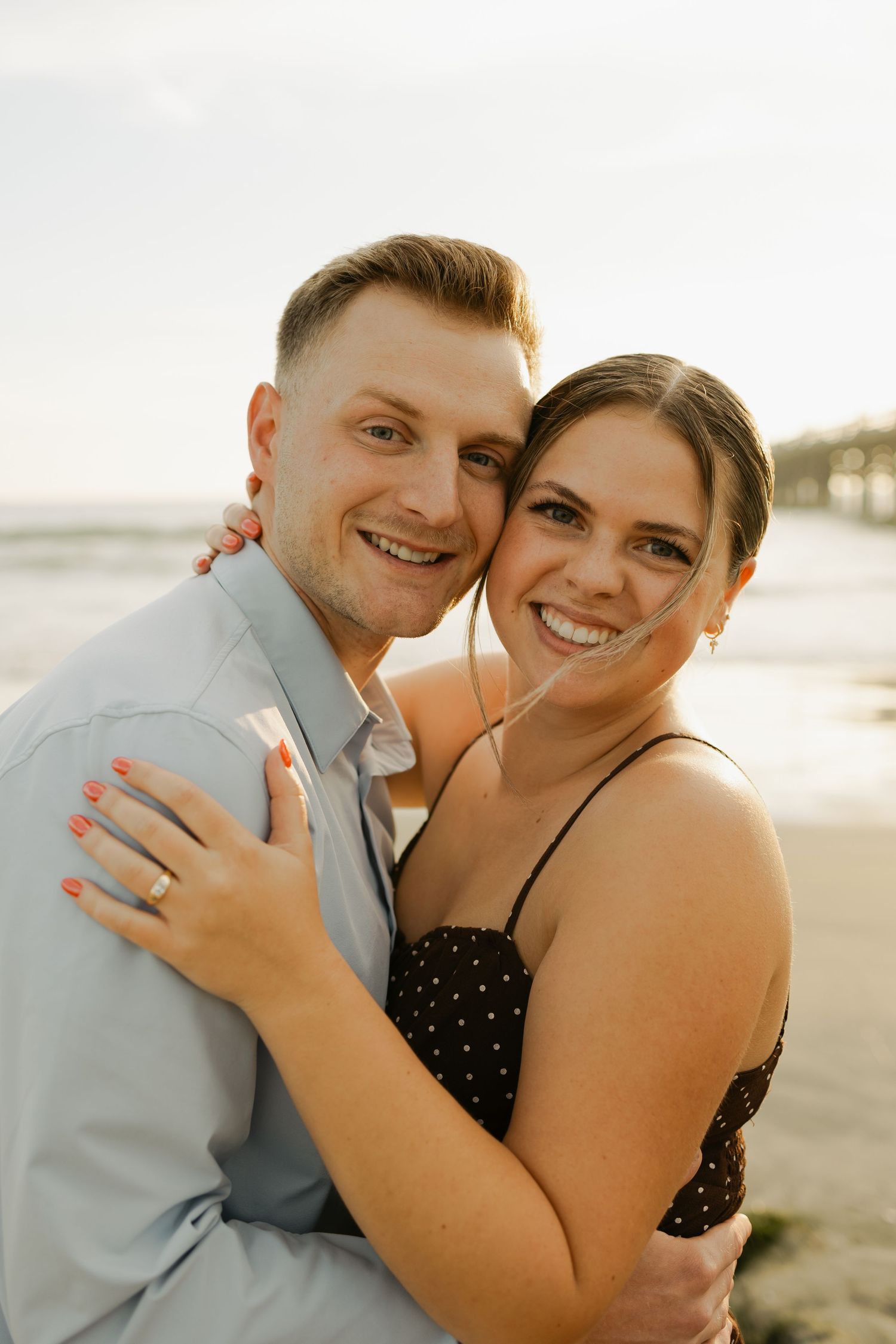 Happy couple embracing on beach at sunset showing off engagement ring.
