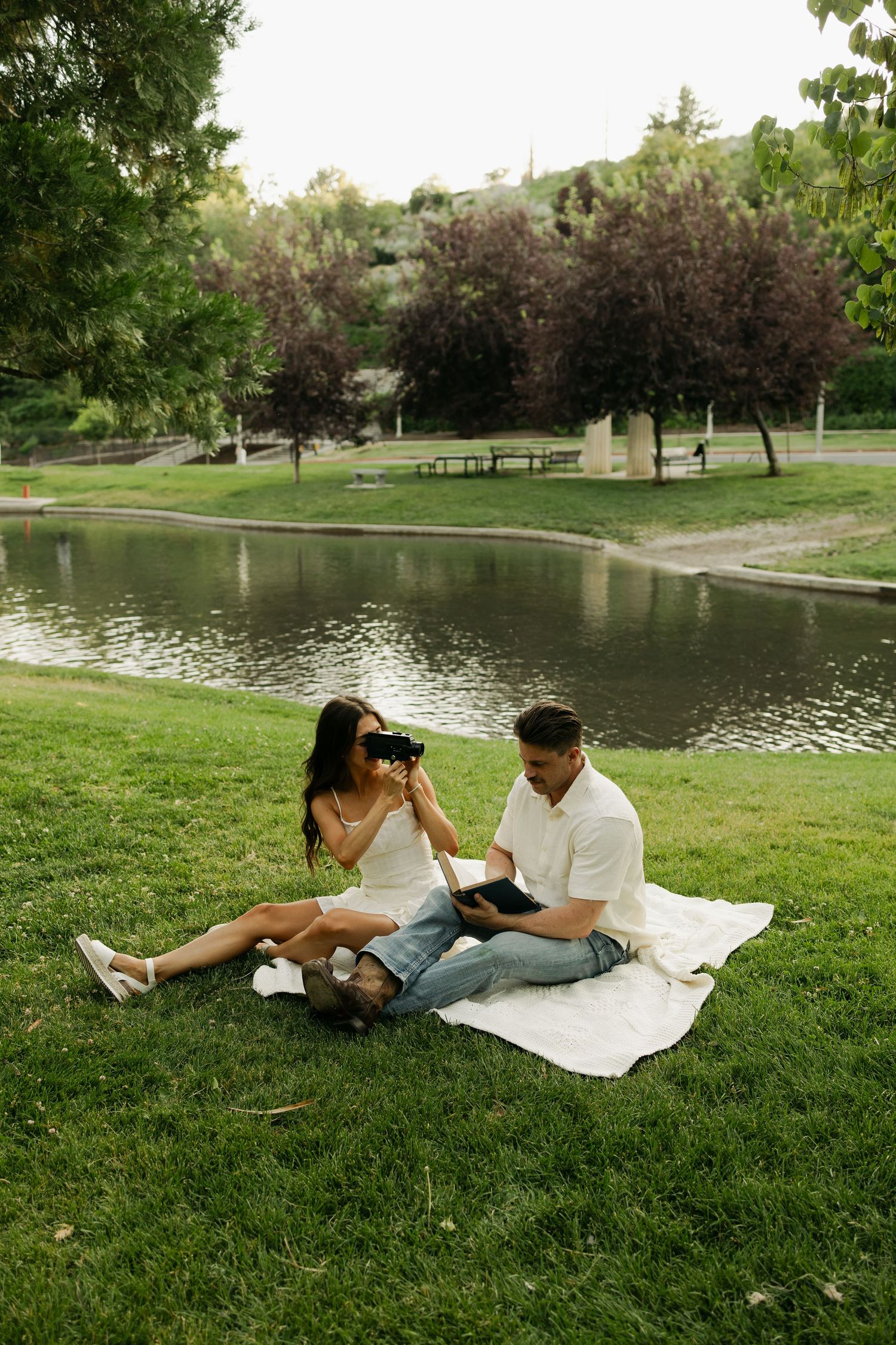 A couple enjoys a romantic picnic on a blanket by a serene pond surrounded by trees in a peaceful park setting.