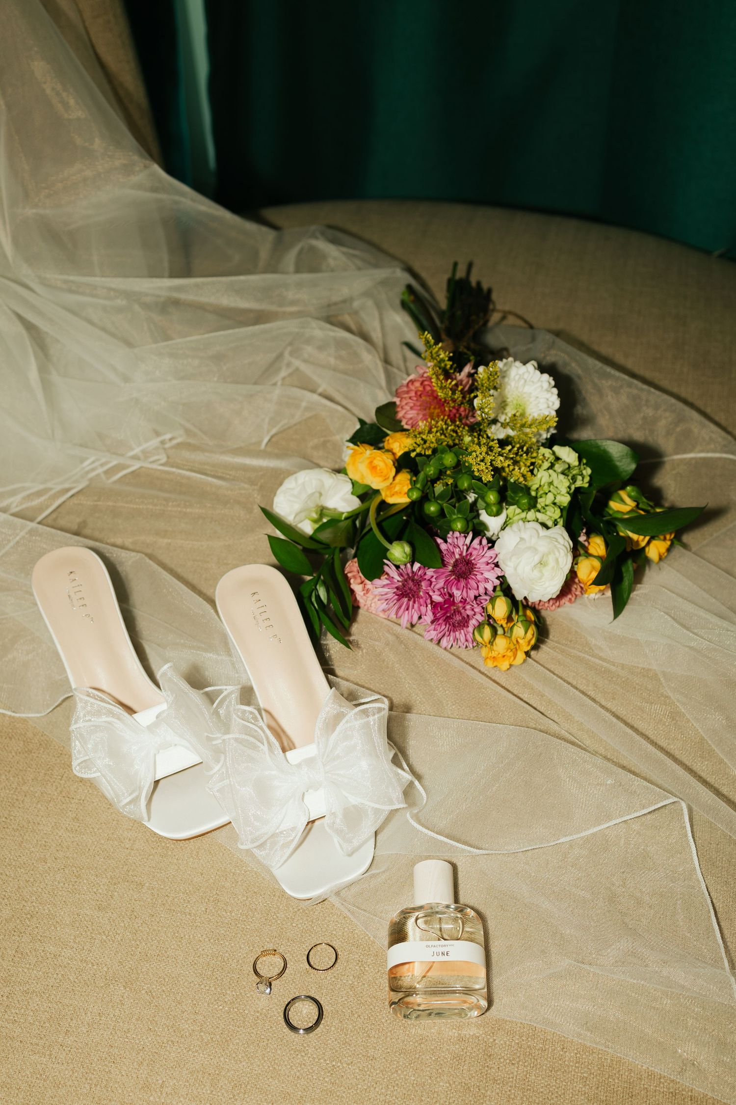 Wedding accessories including white lace heels, wedding rings, perfume bottle and colorful floral bouquet arranged on beige fabric.