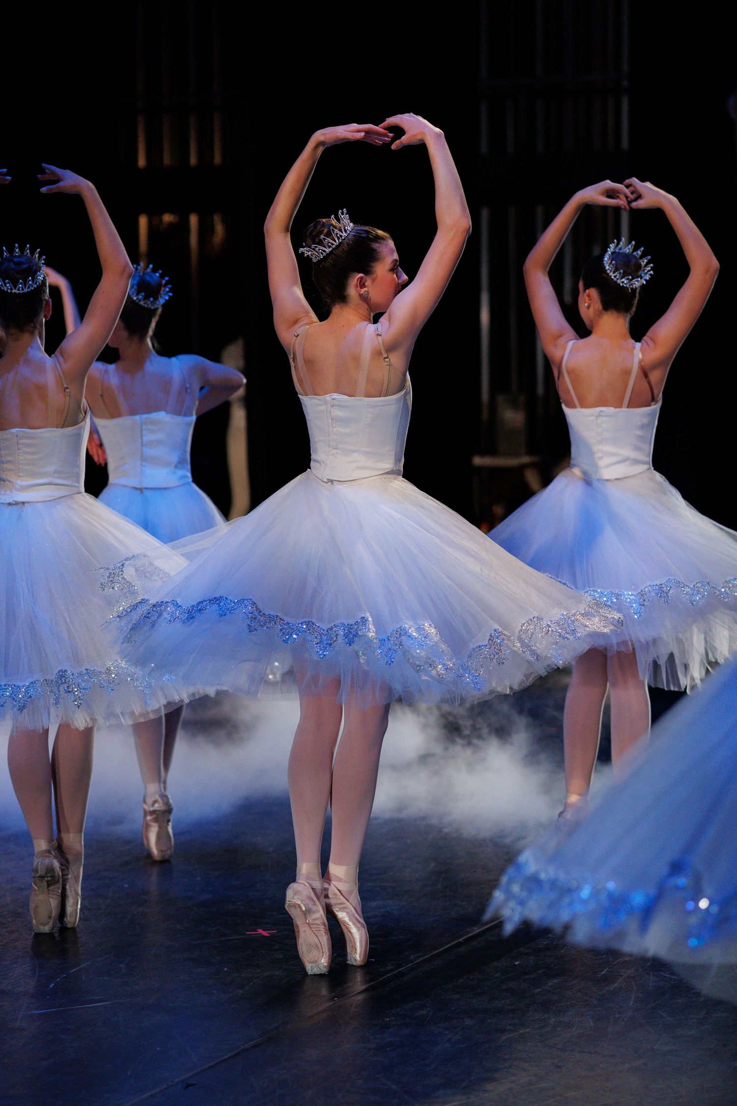 Ballet dancers in white tutus perform en pointe with arms gracefully raised in classical formation.