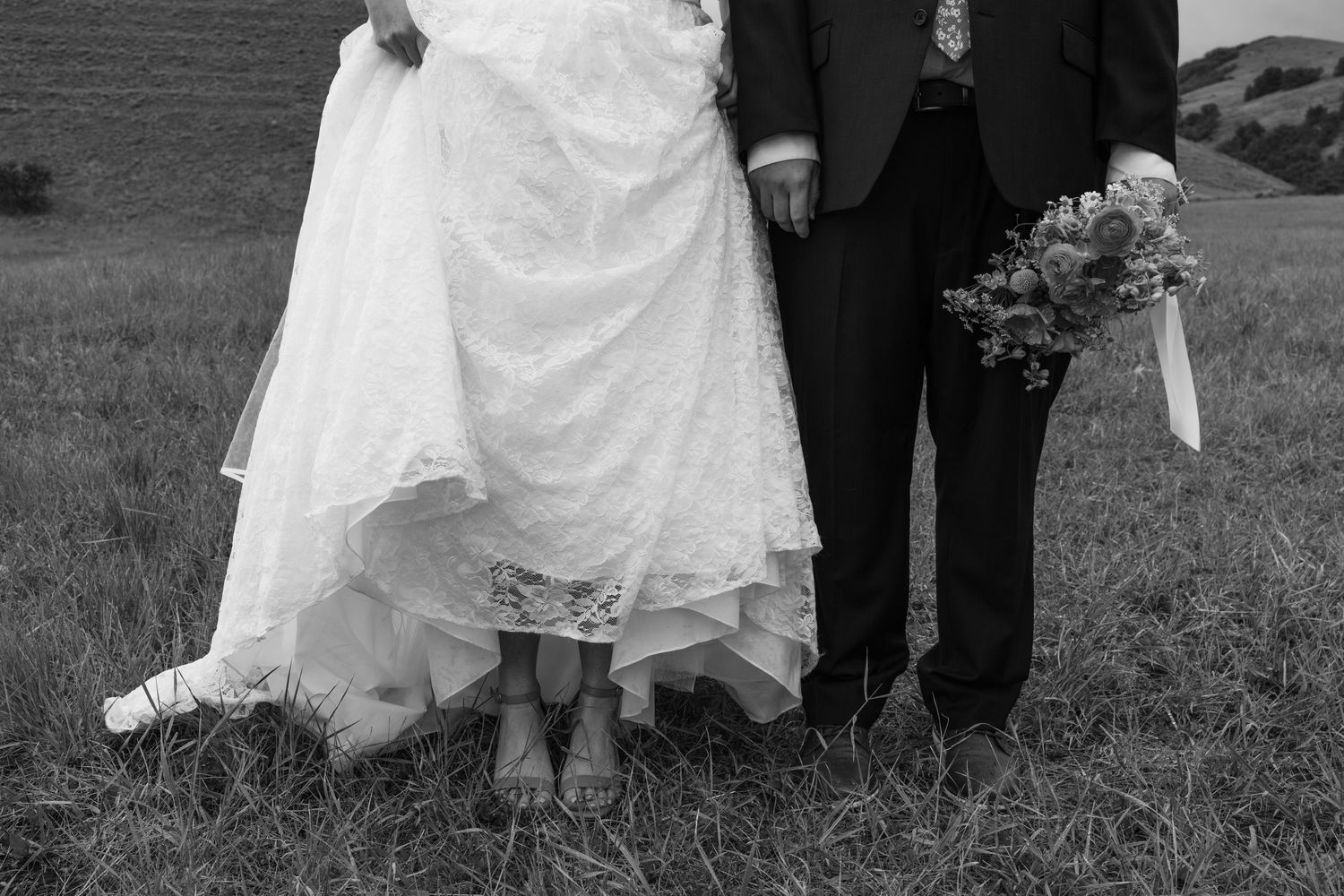 Black and white photo of bride and groom standing in a field, showing wedding dress, suit, and bridal bouquet.
