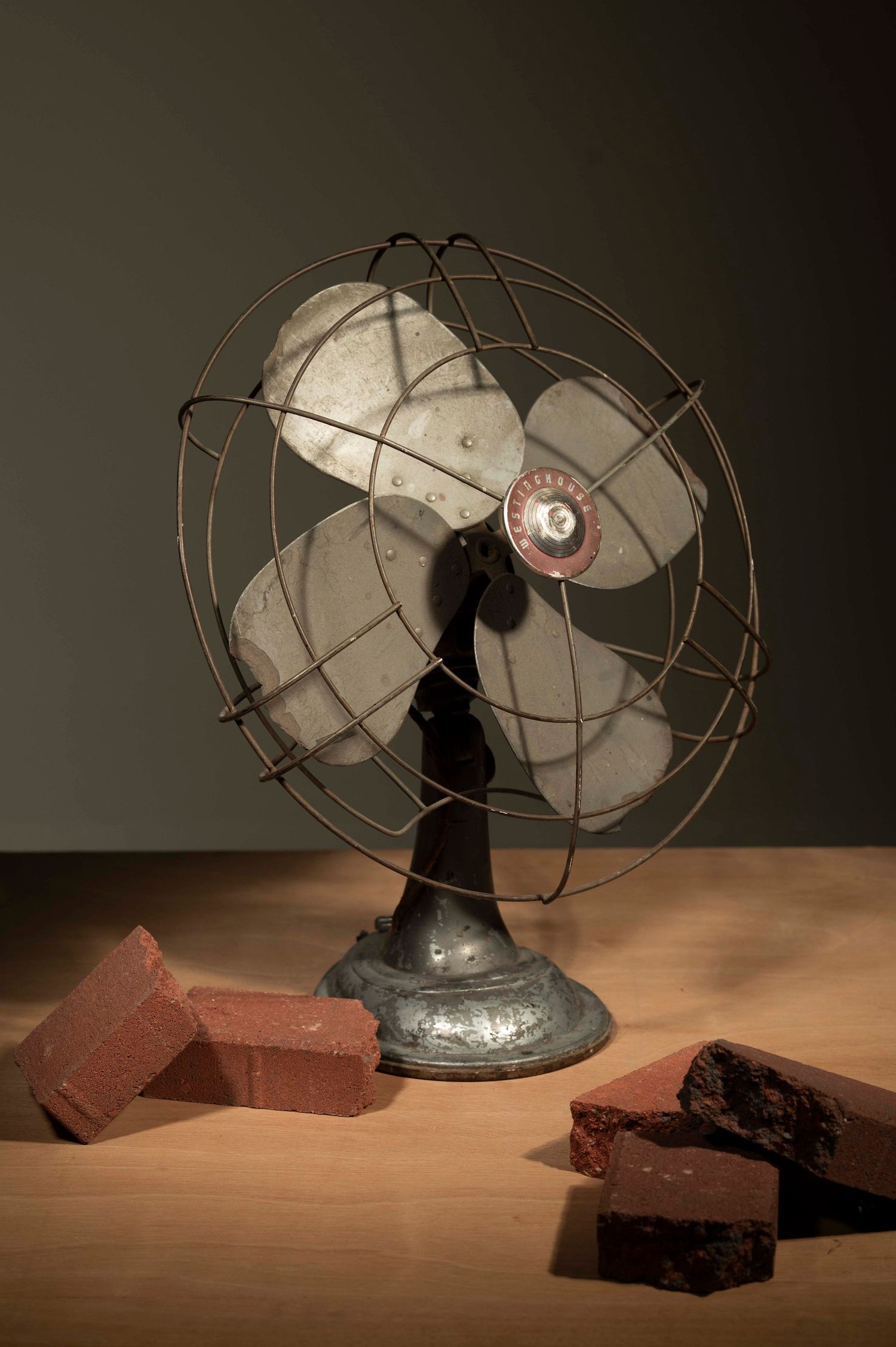 A vintage electric fan sits on a wooden surface beside red bricks in a dark, moody composition.