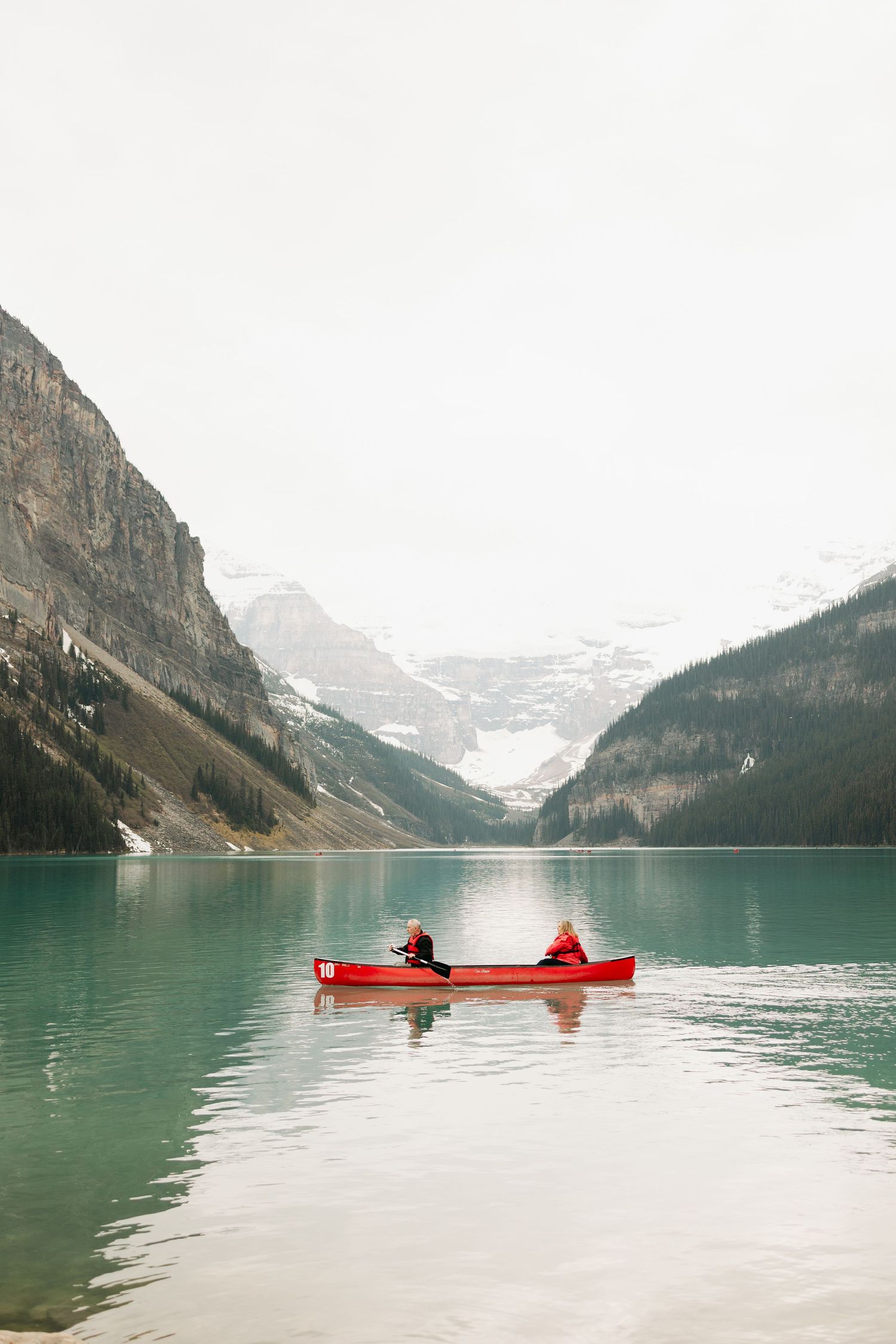 Two people paddling a red canoe on turquoise Lake Louise surrounded by snow-capped mountains in Banff, Canada.
