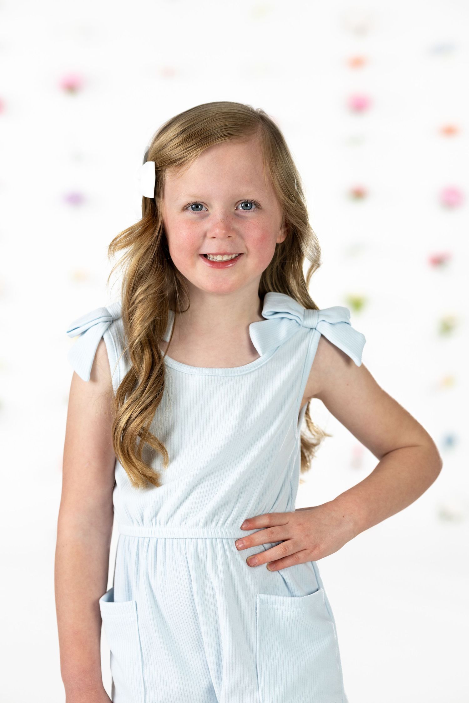Young child in white sleeveless dress poses playfully against floral background with blurred bokeh effect.