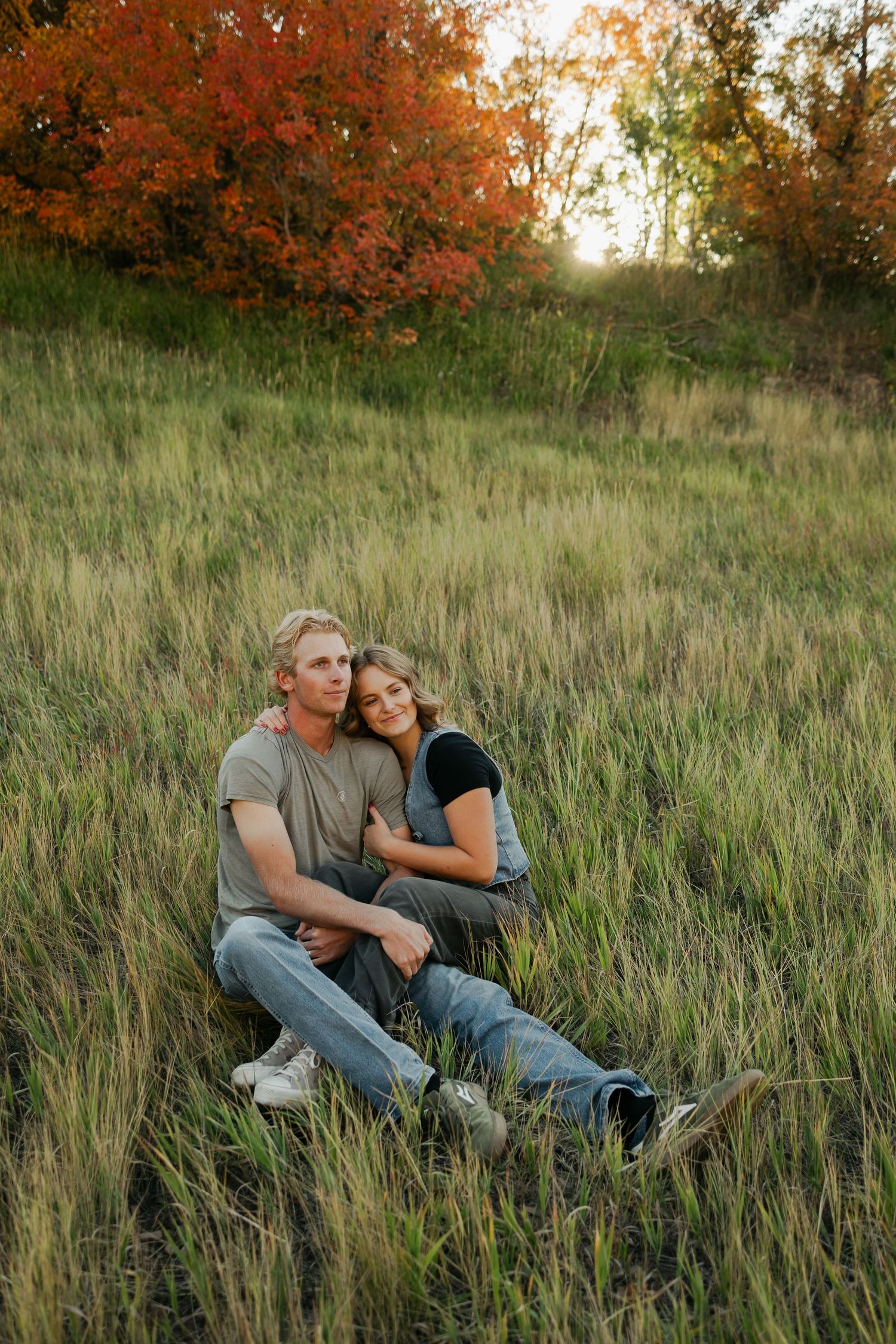 Couple sitting in tall grass with autumn foliage and sunset lighting.