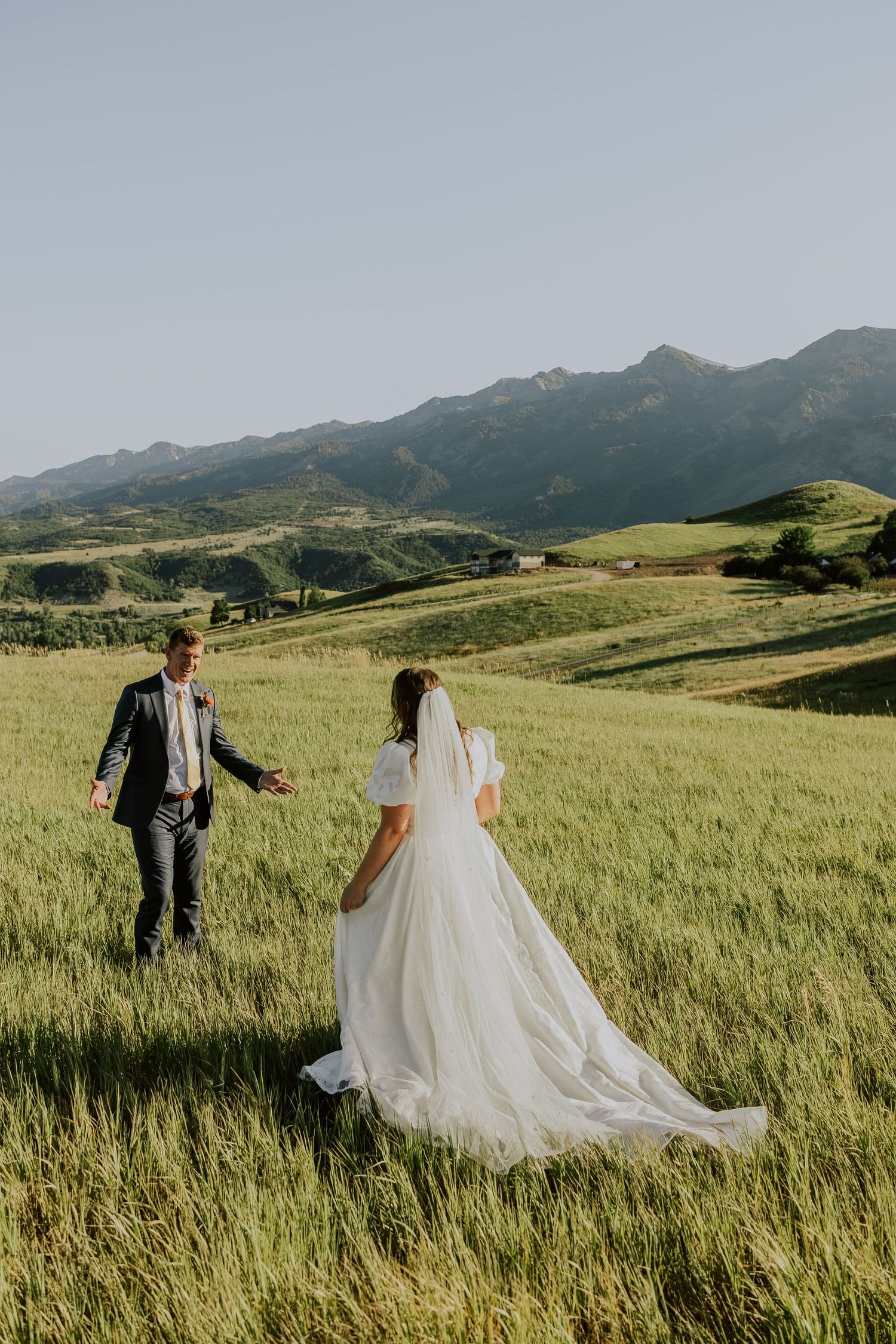 A pair stands together in flowing wedding attire against a lush mountainous backdrop with green rolling hills.
