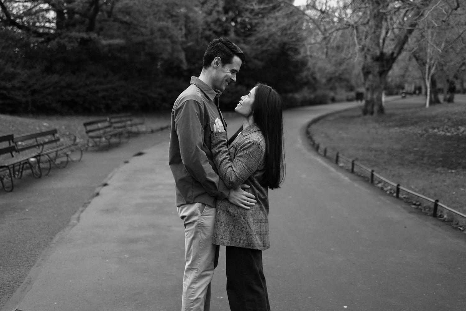 Black and white photo of a couple embracing and almost kissing in a park pathway surrounded by trees.