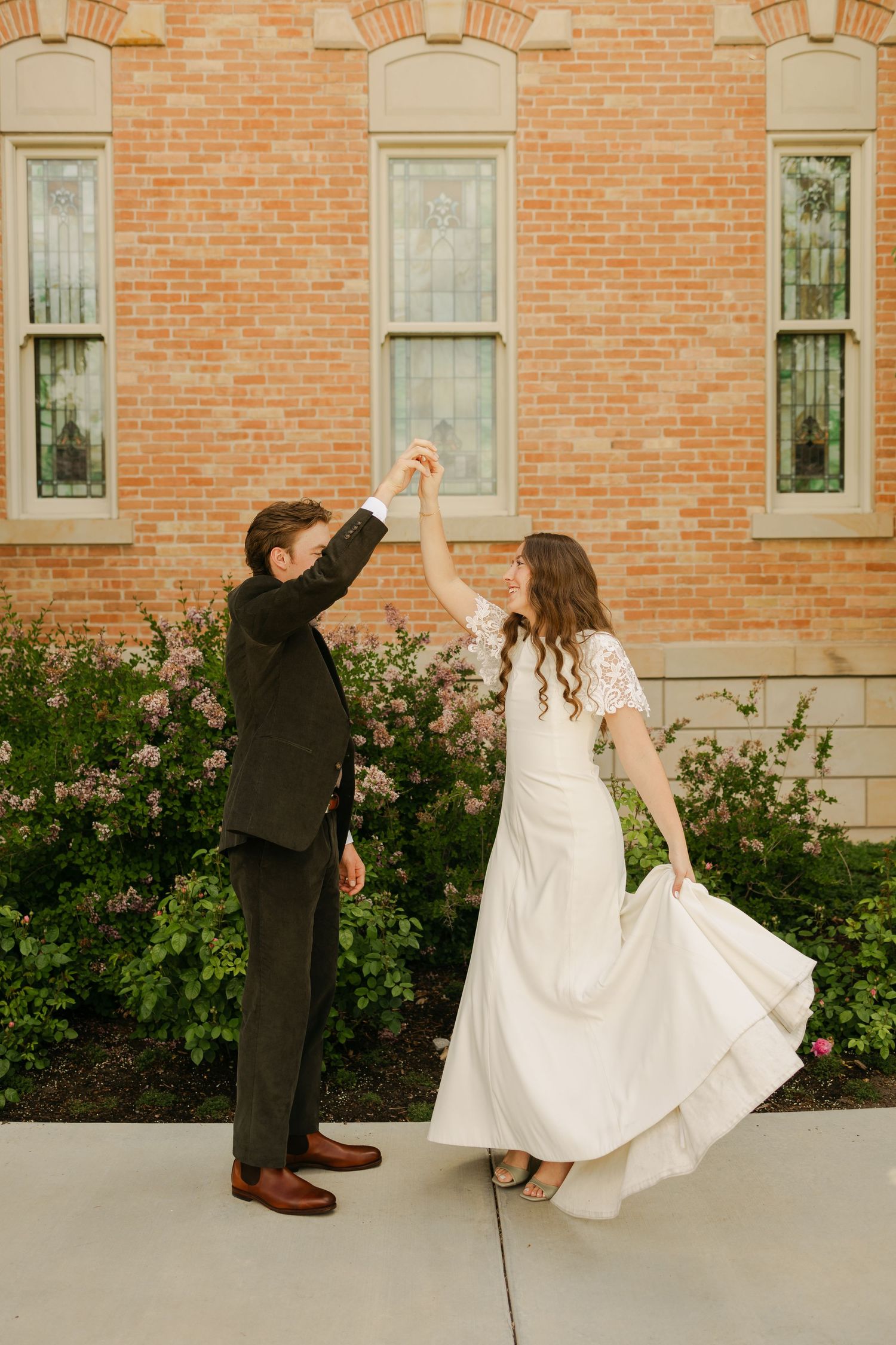 Couple in wedding attire dancing playfully outside brick building.