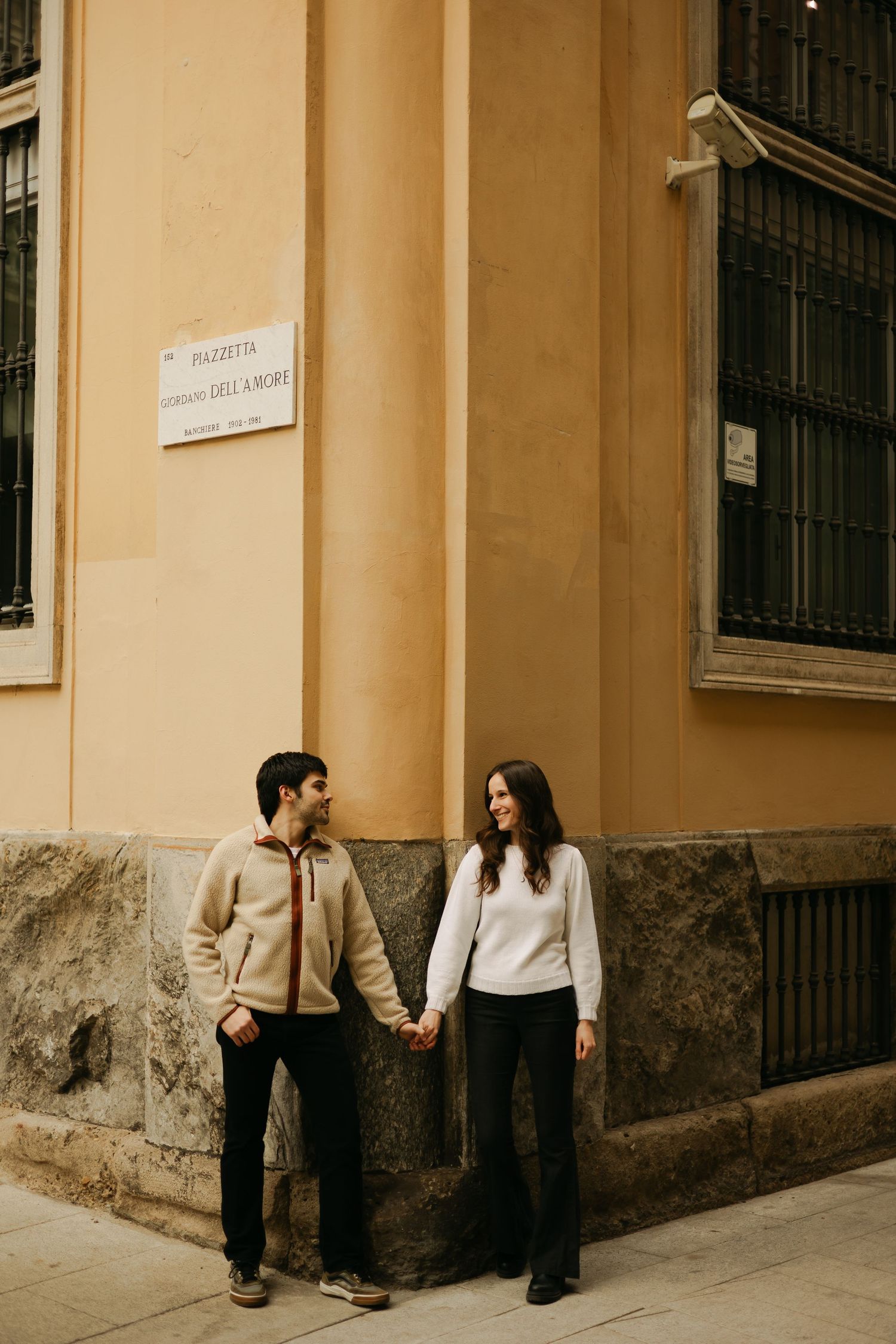 Couple holding hands while standing at corner of historic building in casual attire.