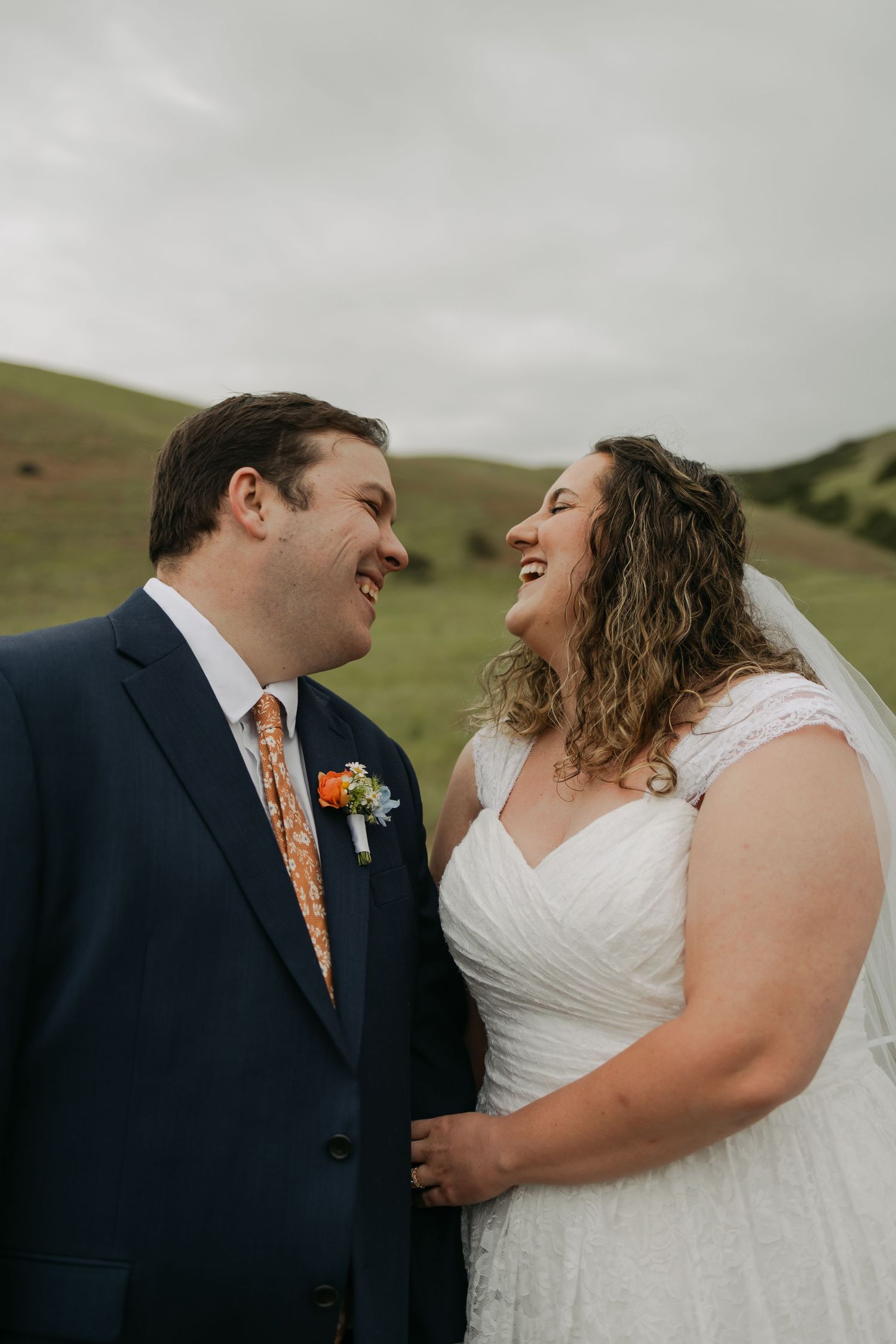 Laughing bride in white gown and groom in navy suit with orange tie share a joyful moment outdoors on their wedding day.