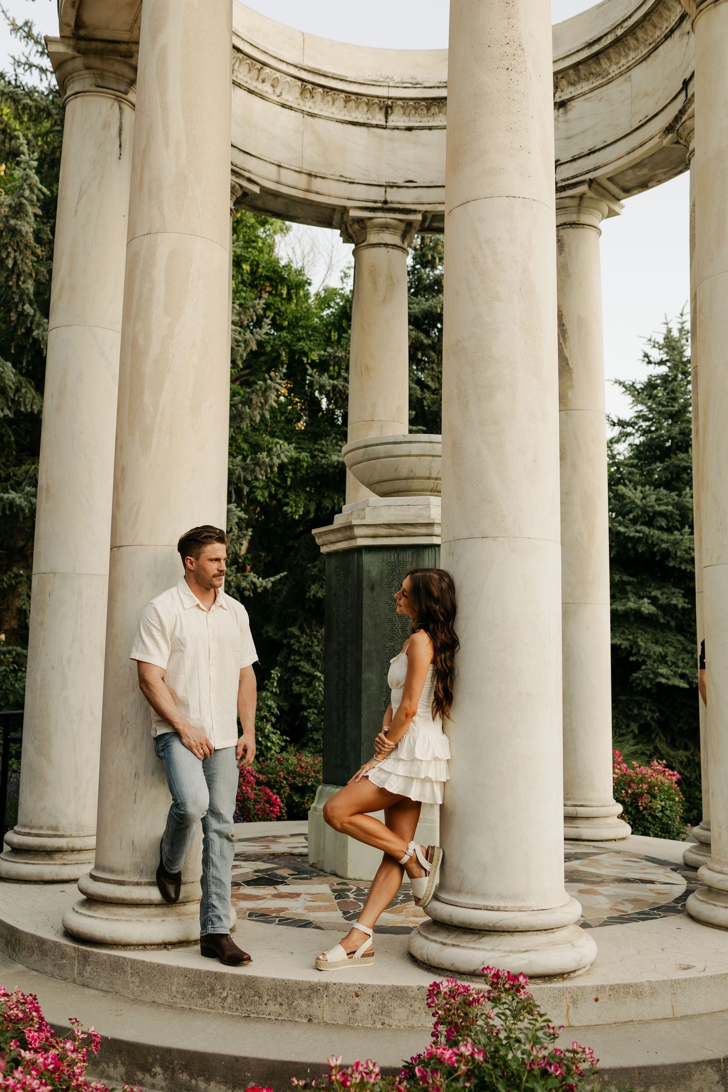 A couple poses in a classical stone gazebo with tall columns surrounded by pink flowers and evergreen trees.