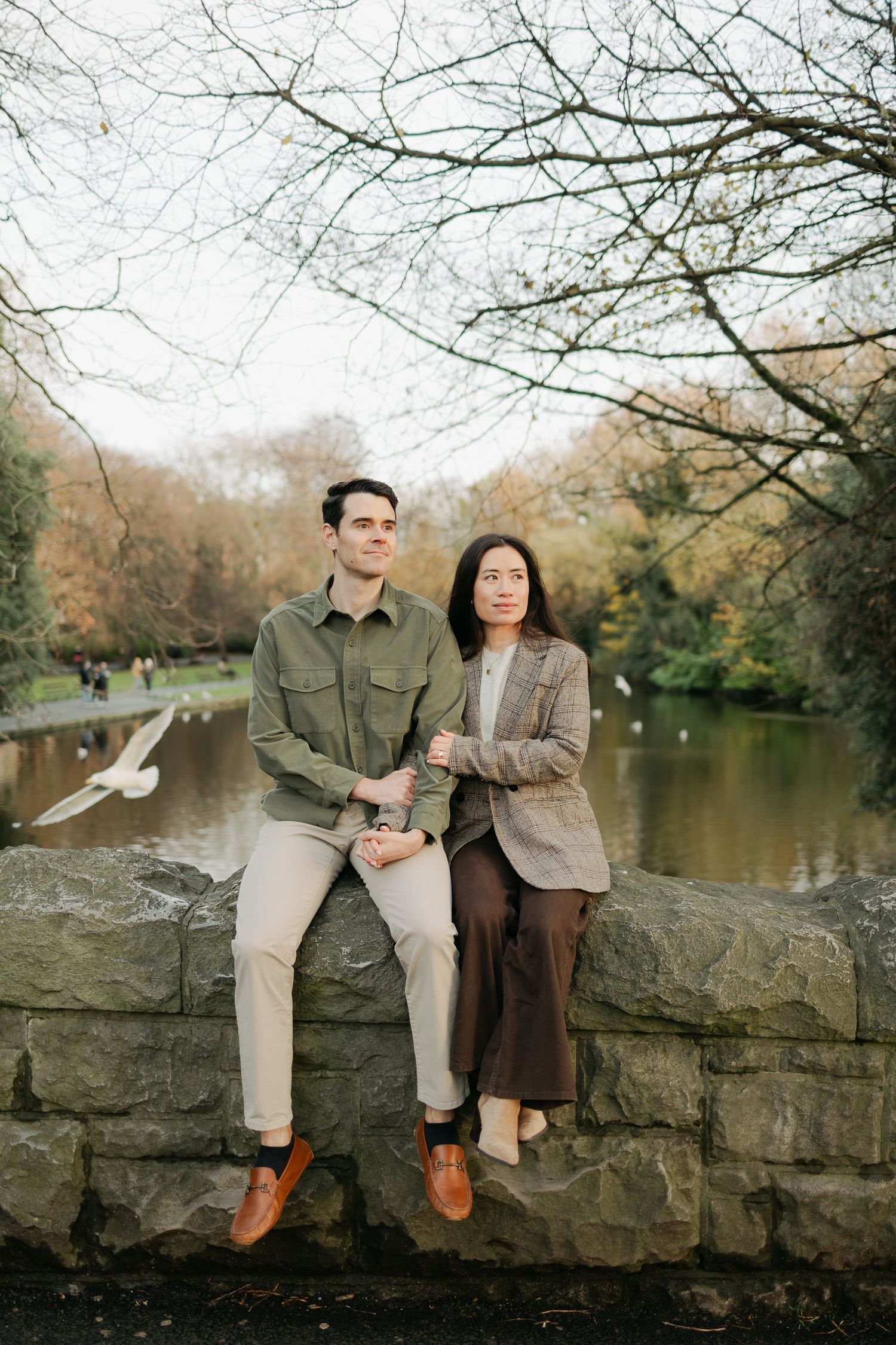 Smiling couple sitting on stone wall by a tranquil park pond surrounded by autumn foliage and flying birds.