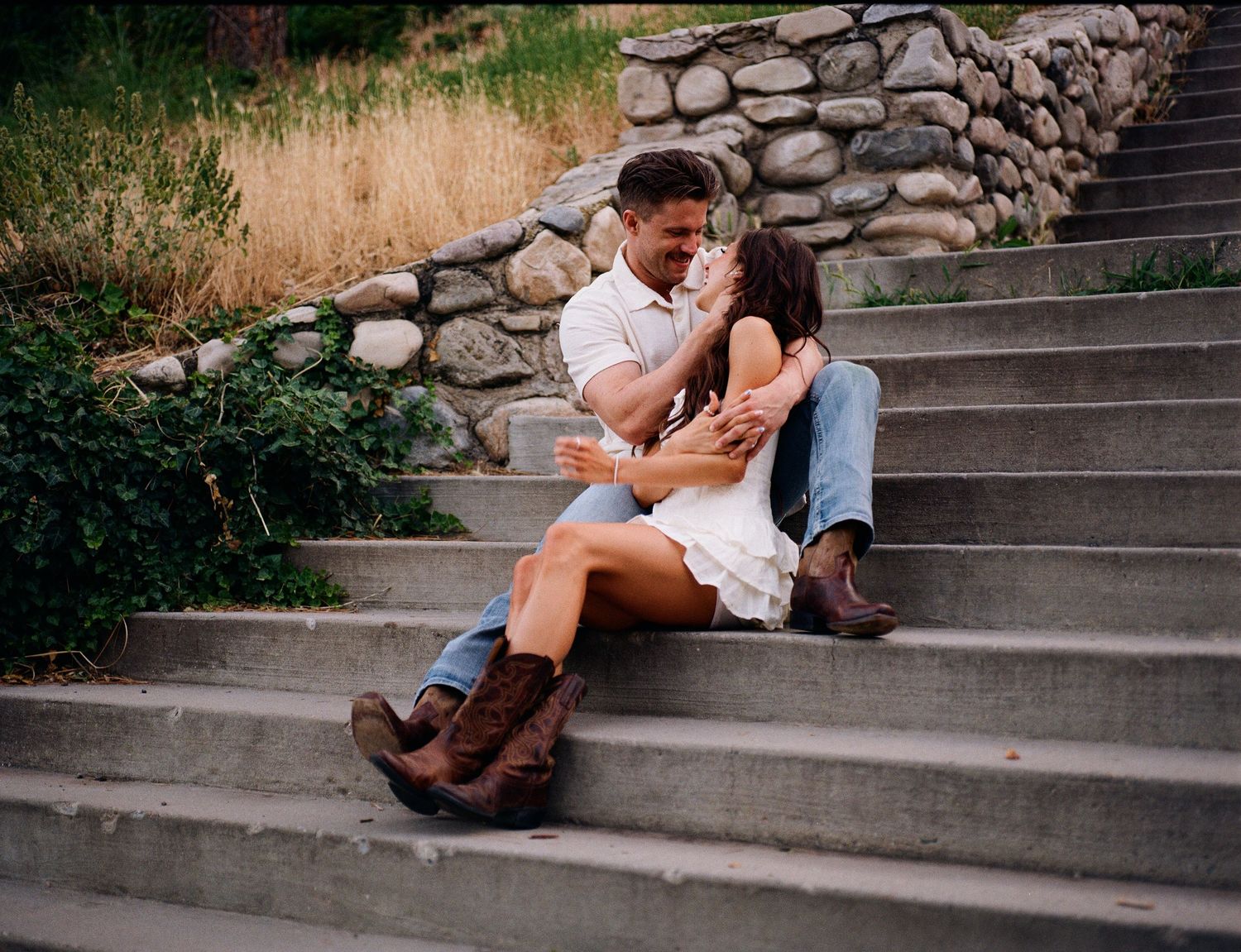 A couple sits intimately on stone steps against a rustic wall on a warm summer evening.