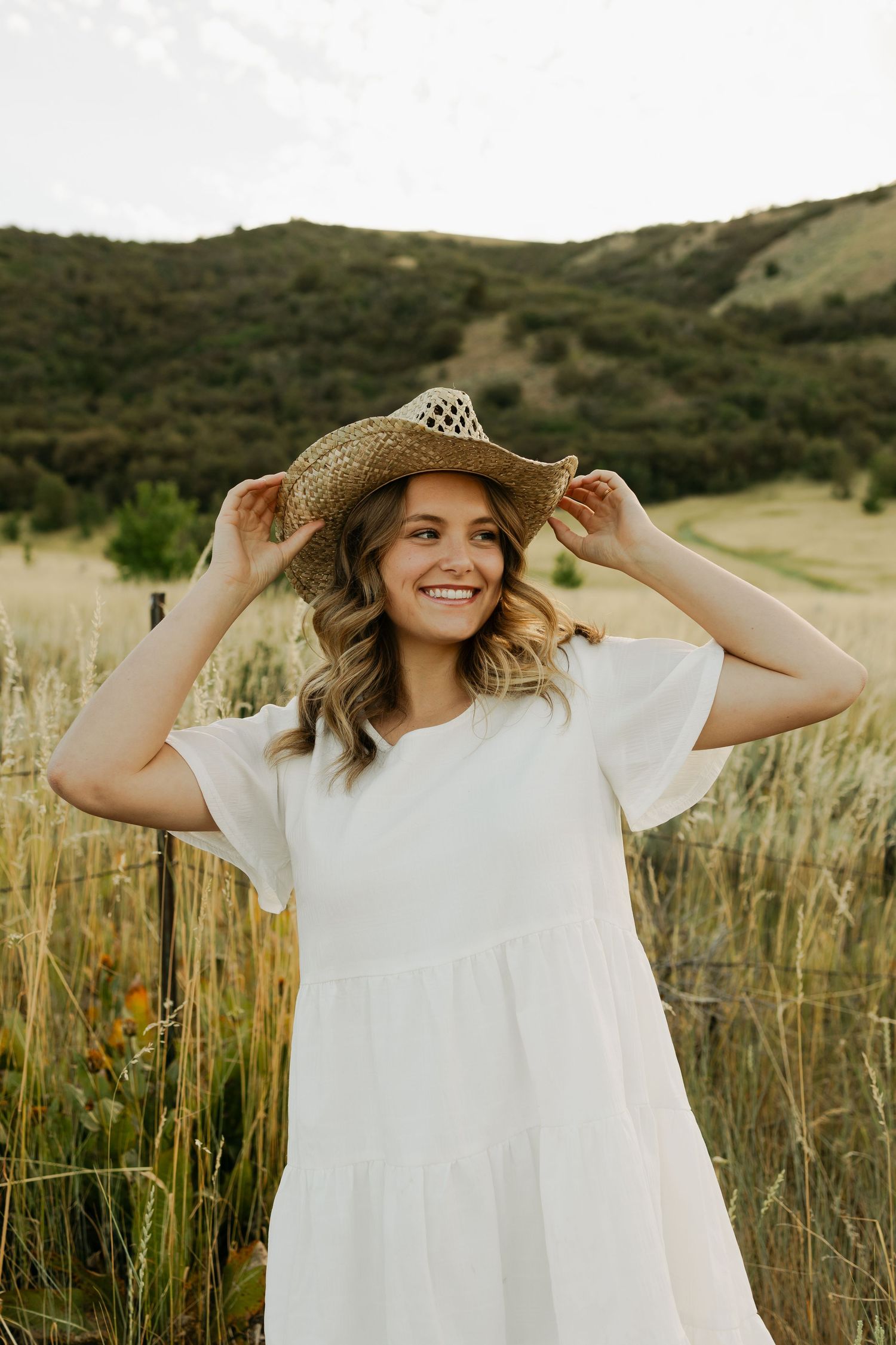 Person in white dress adjusting straw hat in golden field with mountains in background.