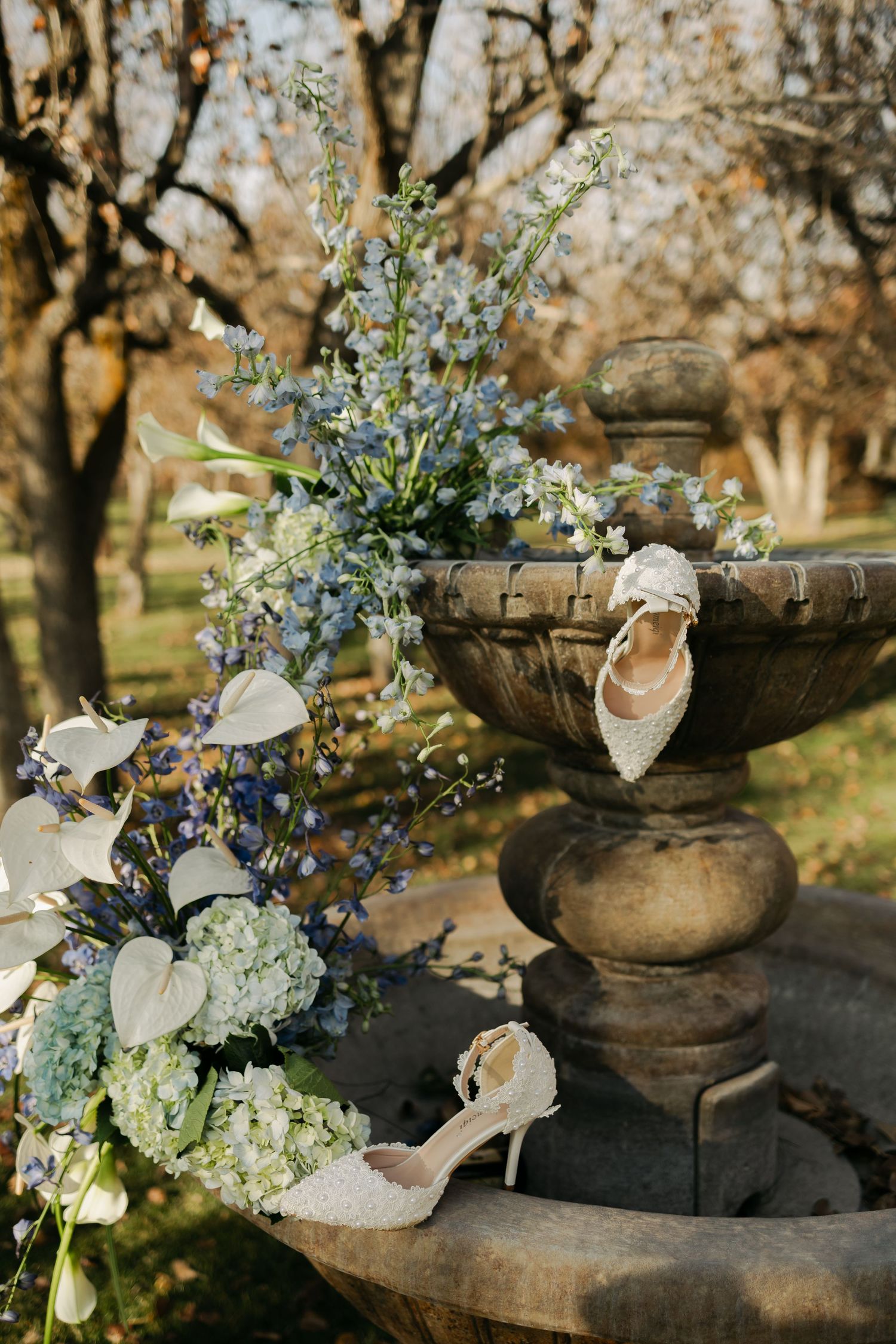 Elegant wedding shoes and floral arrangement displayed on an ornate stone fountain in a garden setting.