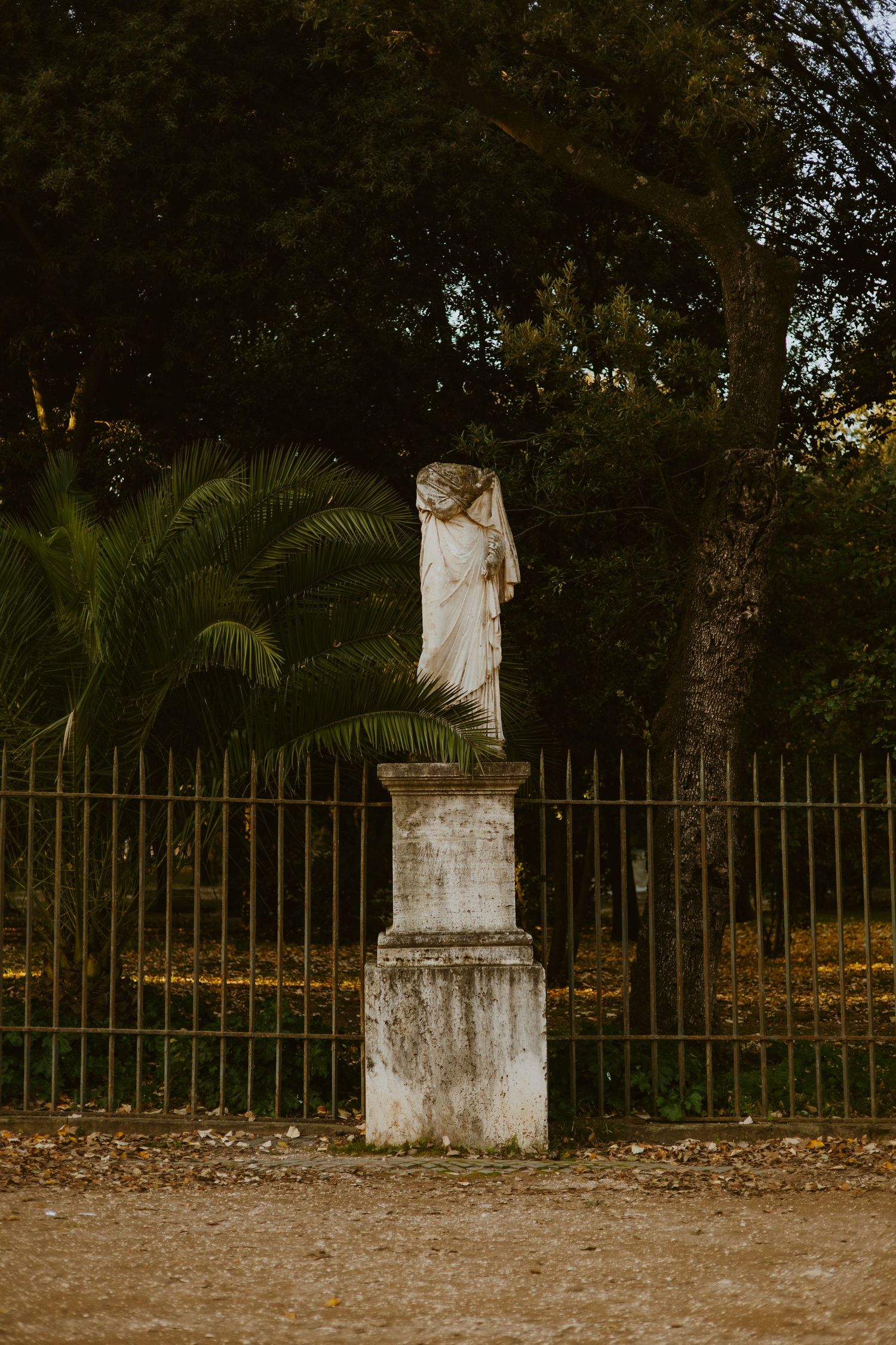 Ancient headless marble statue on stone pedestal beside iron fence, surrounded by palm trees in a garden.