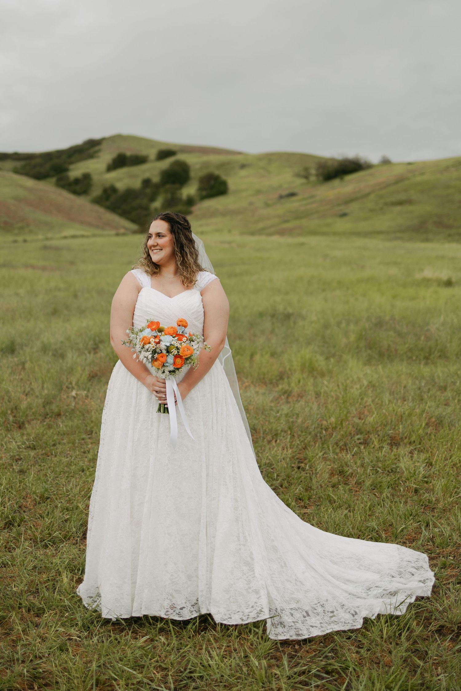 Bride in white lace gown holding orange bouquet stands in green rolling hills landscape.