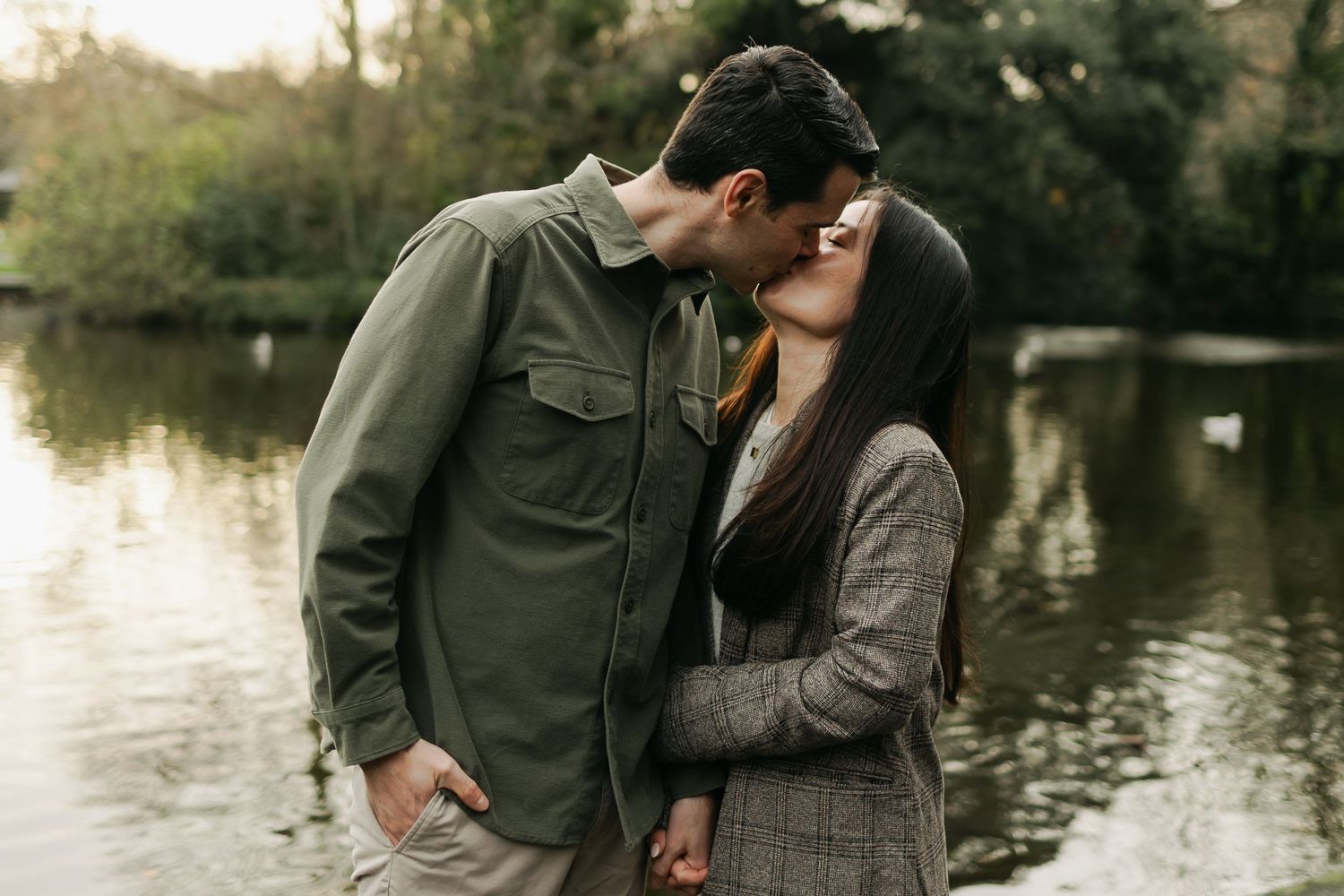 Romantic couple sharing a kiss by a serene lake surrounded by lush greenery in a park.