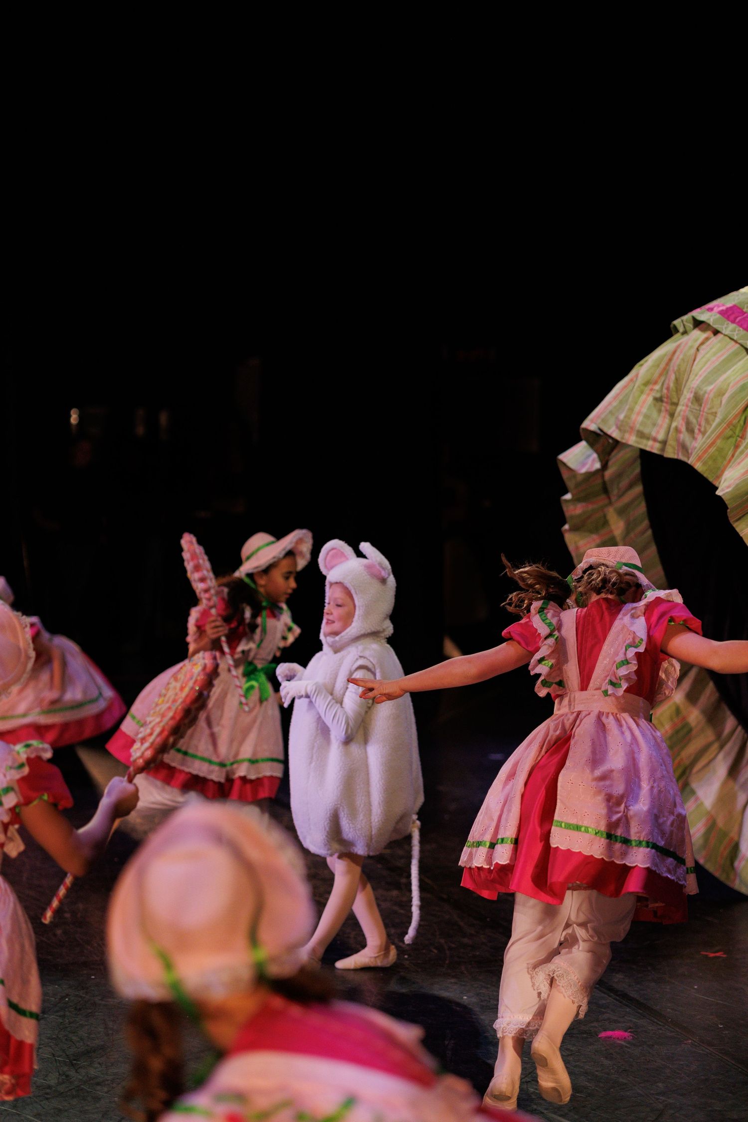 Performers in pink and white costumes dance together on a dark stage during what appears to be a theatrical production.