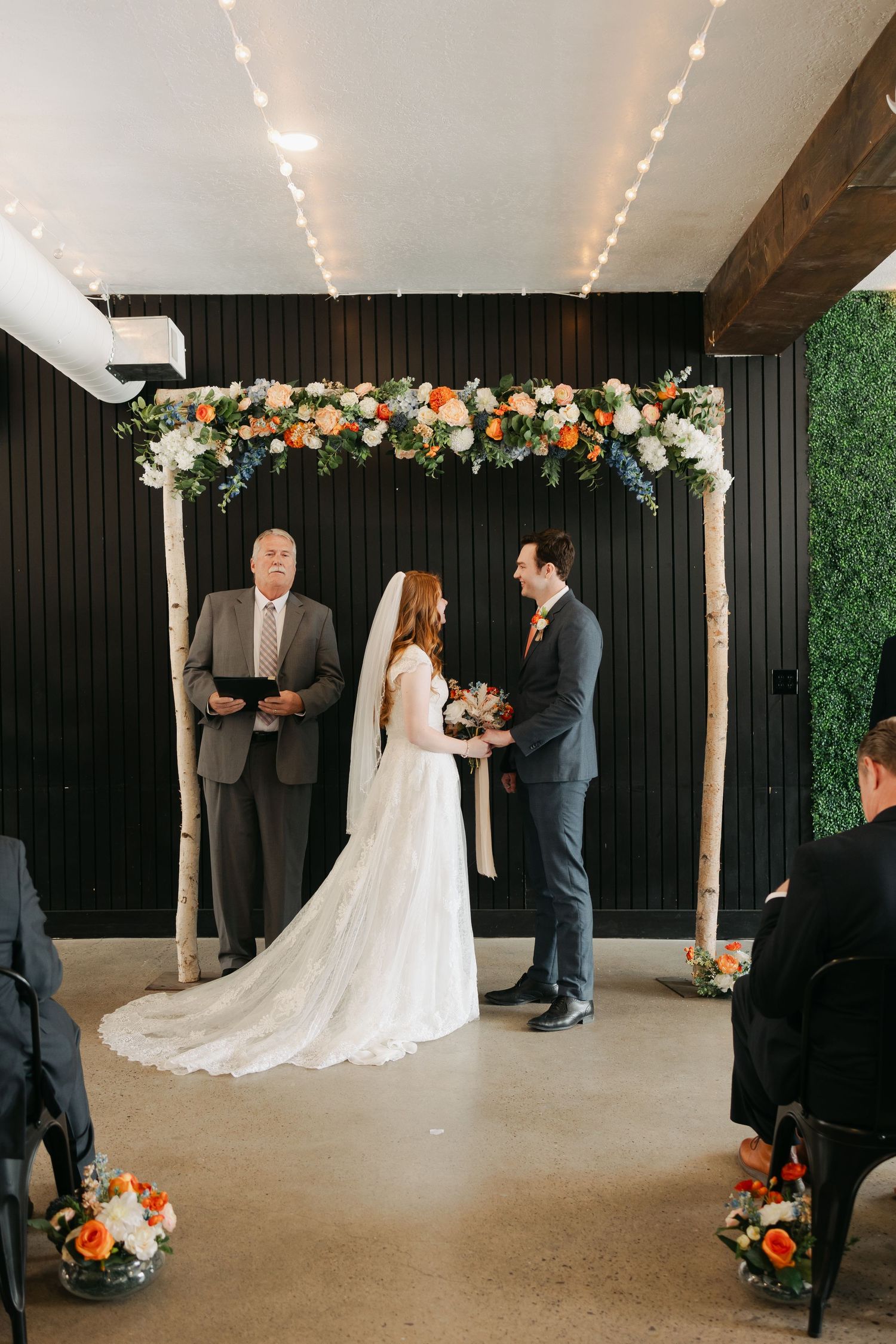 An indoor wedding ceremony with floral arch decorations and string lights overhead.